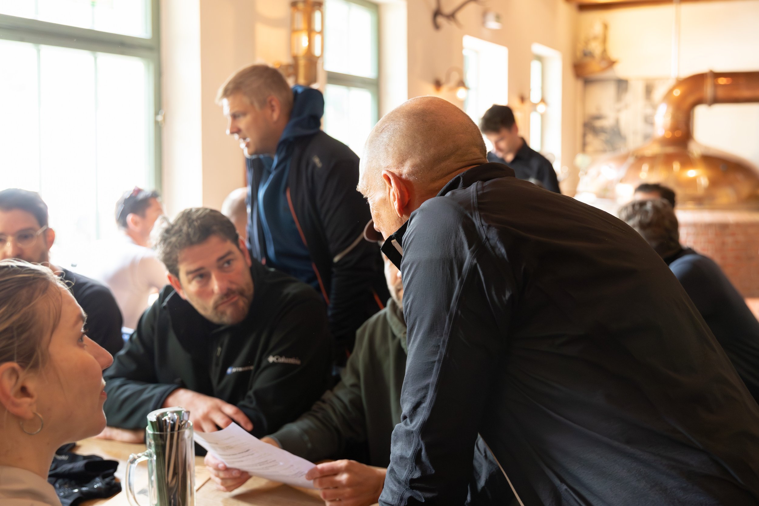 Mehrere Menschen sitzen an einem Tisch in einem rustikalen Restaurant, während ein Mann mit Glatze und schwarzen Jacke eine Unterlage zeigt, um eine Diskussion oder Geschäftsbesprechung zu führen. Im Hintergrund sind große Fenster und eine traditione