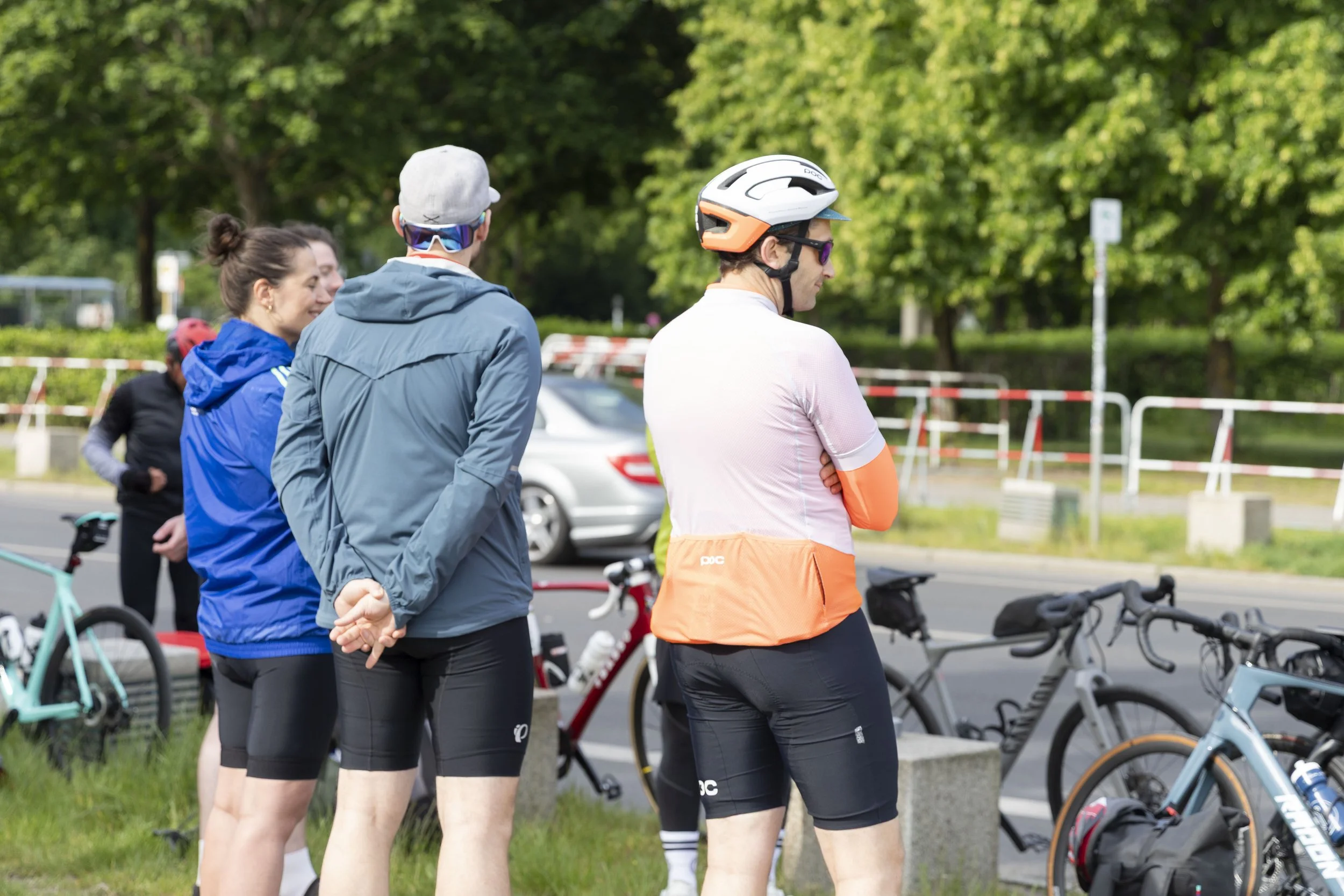 Gruppe von Radfahrern in Fahrradausrüstung steht bei einem Treffen in einer Parkanlage mit Fahrrädern und Bäumen im Hintergrund.