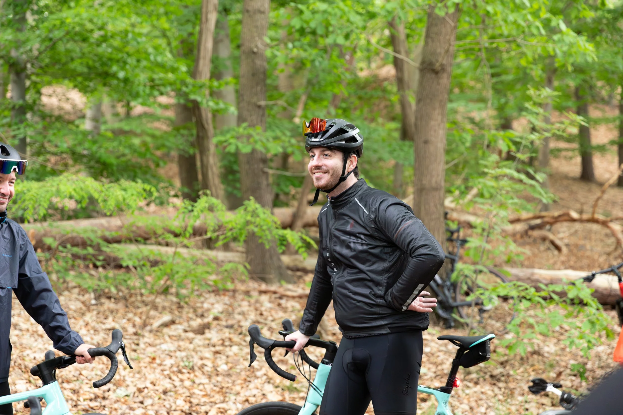 Zwei Radfahrer in einem Wald, einer lächelnd, beide tragen Helme und Radbekleidung, mit Fahrrädern.