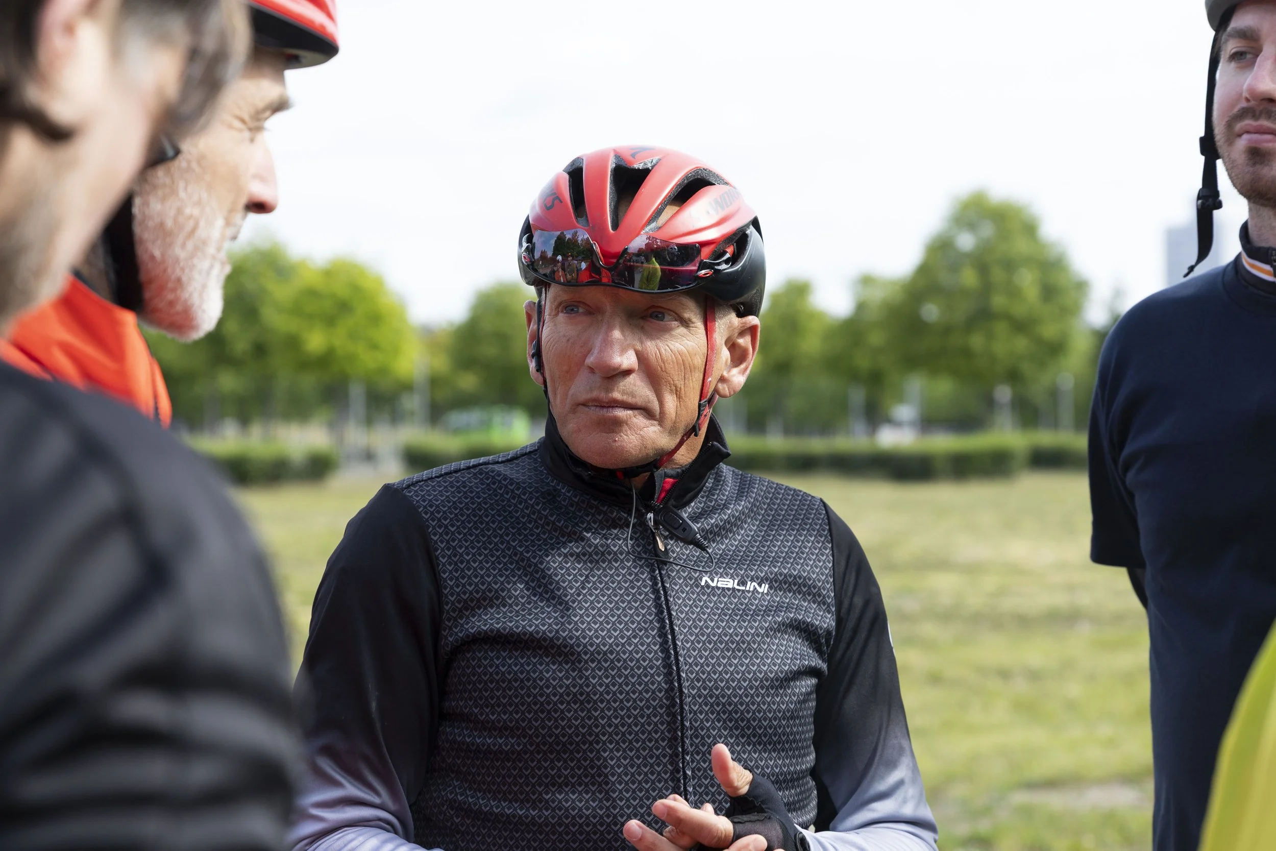 Älterer Mann mit Fahrradhelm und Radsportbekleidung spricht mit drei anderen Personen im Freien in einer grünen Park- oder Wiesenlandschaft.