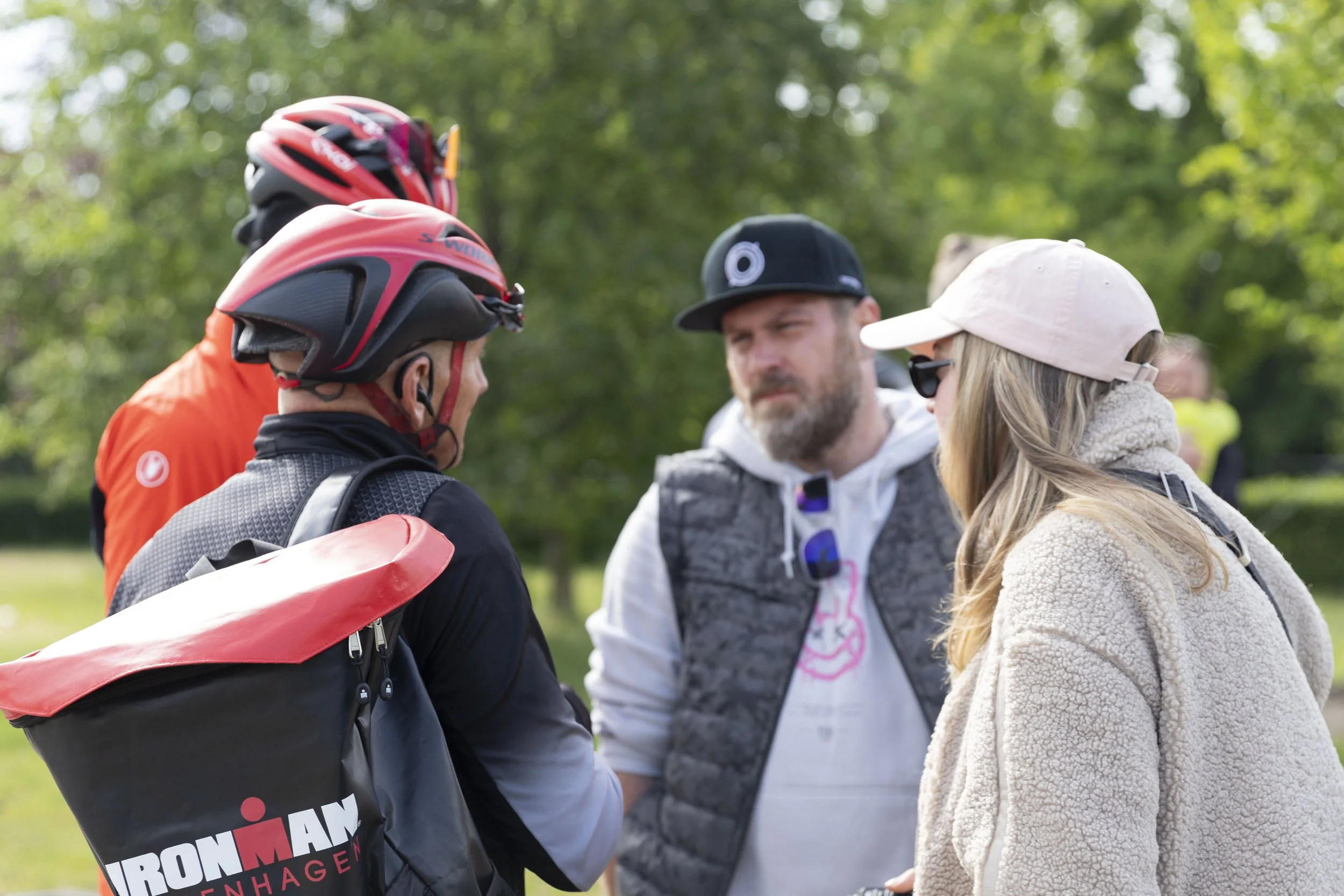 Vier Personen im Gespräch im Freien, zwei Männer in Radhelmen und Outdoor-Kleidung, eine Frau mit Sonnenbrille und Baseballcap, im Hintergrund grüne Bäume.