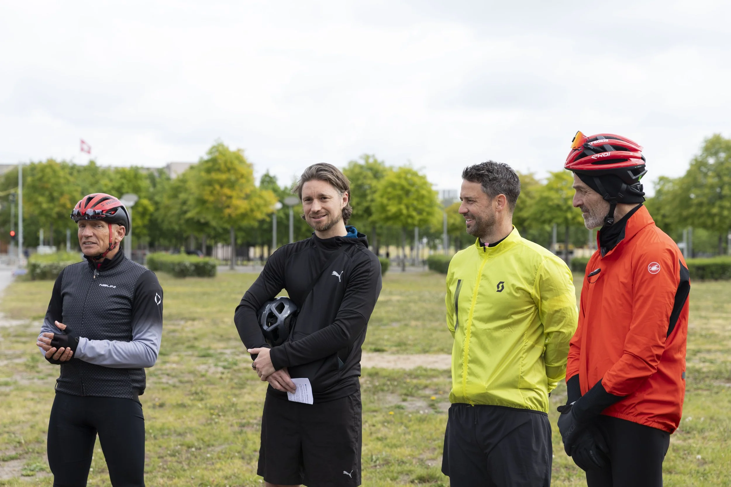 Vier Männer stehen im Freien auf einer Wiese, alle tragen Fahrradhelme und Sportkleidung, vermutlich vor einer Radtour oder einem Training.