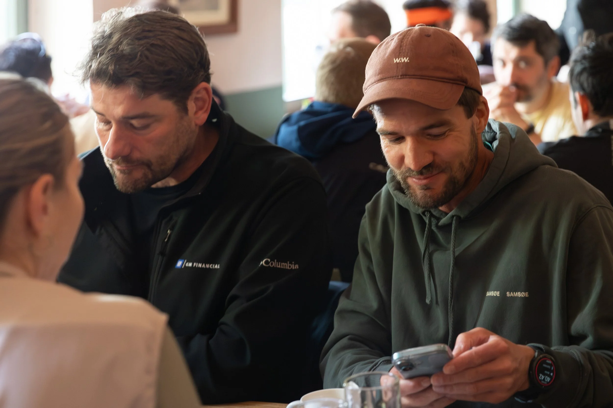 Zwei Männer sitzen in einem Café, einer schaut auf sein Smartphone, der andere hat die Augen geschlossen.