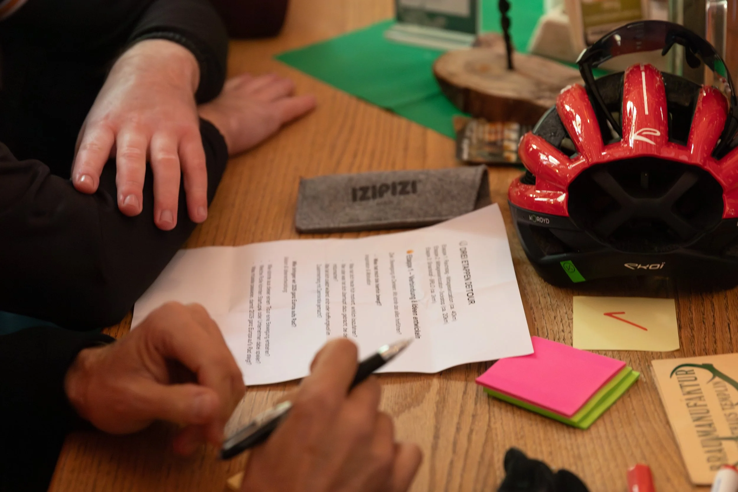 Menschen sitzen an einem Tisch, auf dem Fahrradhelm, Handschuhe, Notizzettel, Redezeichen, ein Schild mit 'IZIPIZI', eine Zeitung und andere Gegenstände liegen.