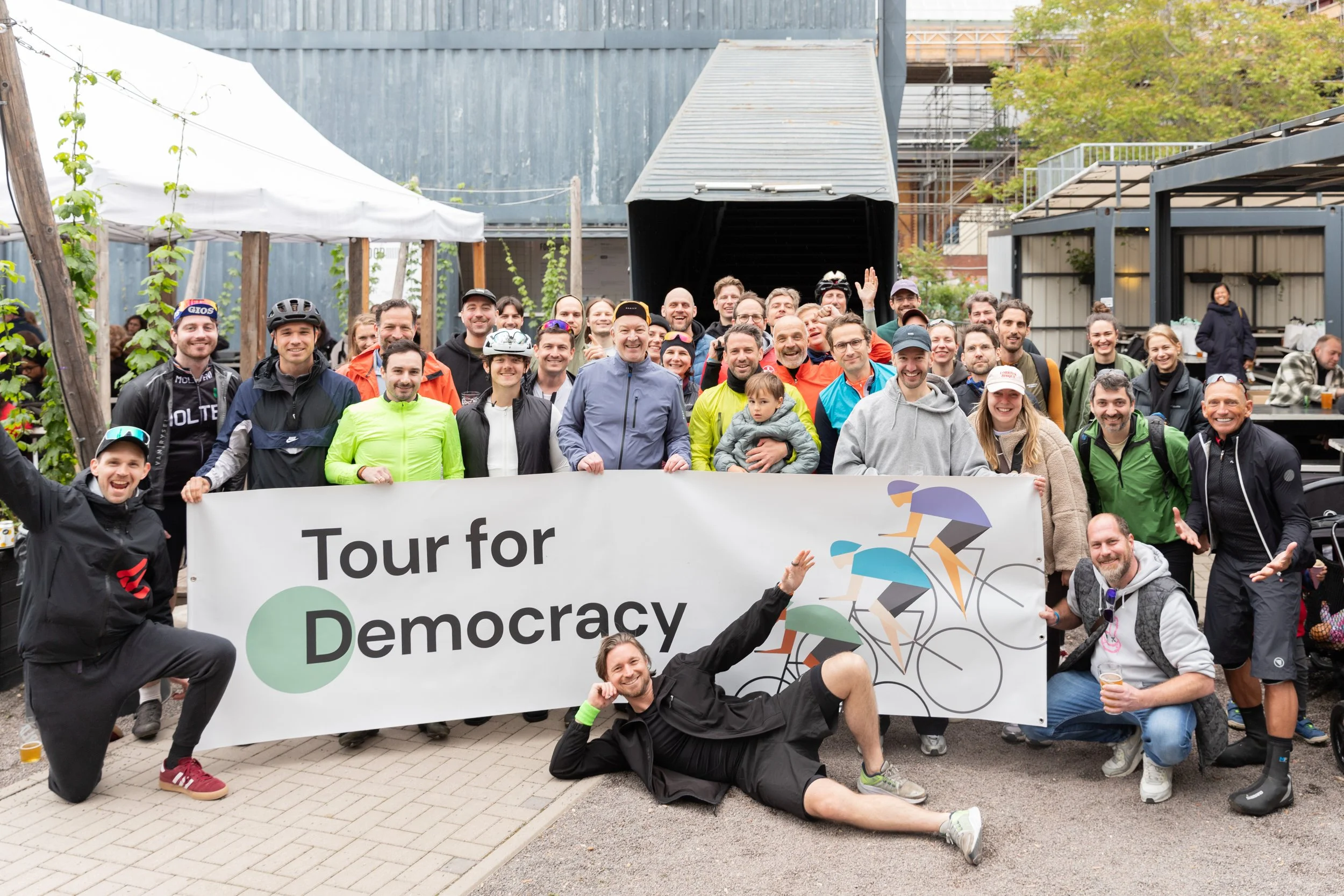Gruppe von Menschen bei einer Fahrradtour, hält ein Banner mit der Aufschrift 'Tour for Democracy', im Hintergrund sind Bäume und ein Gebäude.