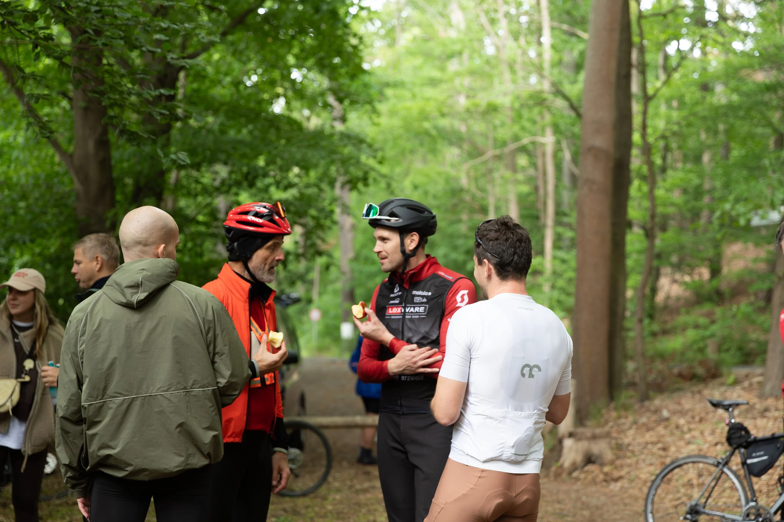 Gruppe von Menschen, darunter Radfahrer in Helm, im Gespräch in einem Wald, während ein Fahrrad im Hintergrund steht, mit grünen Bäumen.