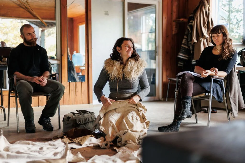 Trois personnes assises dans une pièce en bois, échangeant probablement lors d'une réunion ou d'un atelier. La femme au centre est souriante, la femme à droite prend des notes, et l'homme à gauche écoute attentivement.