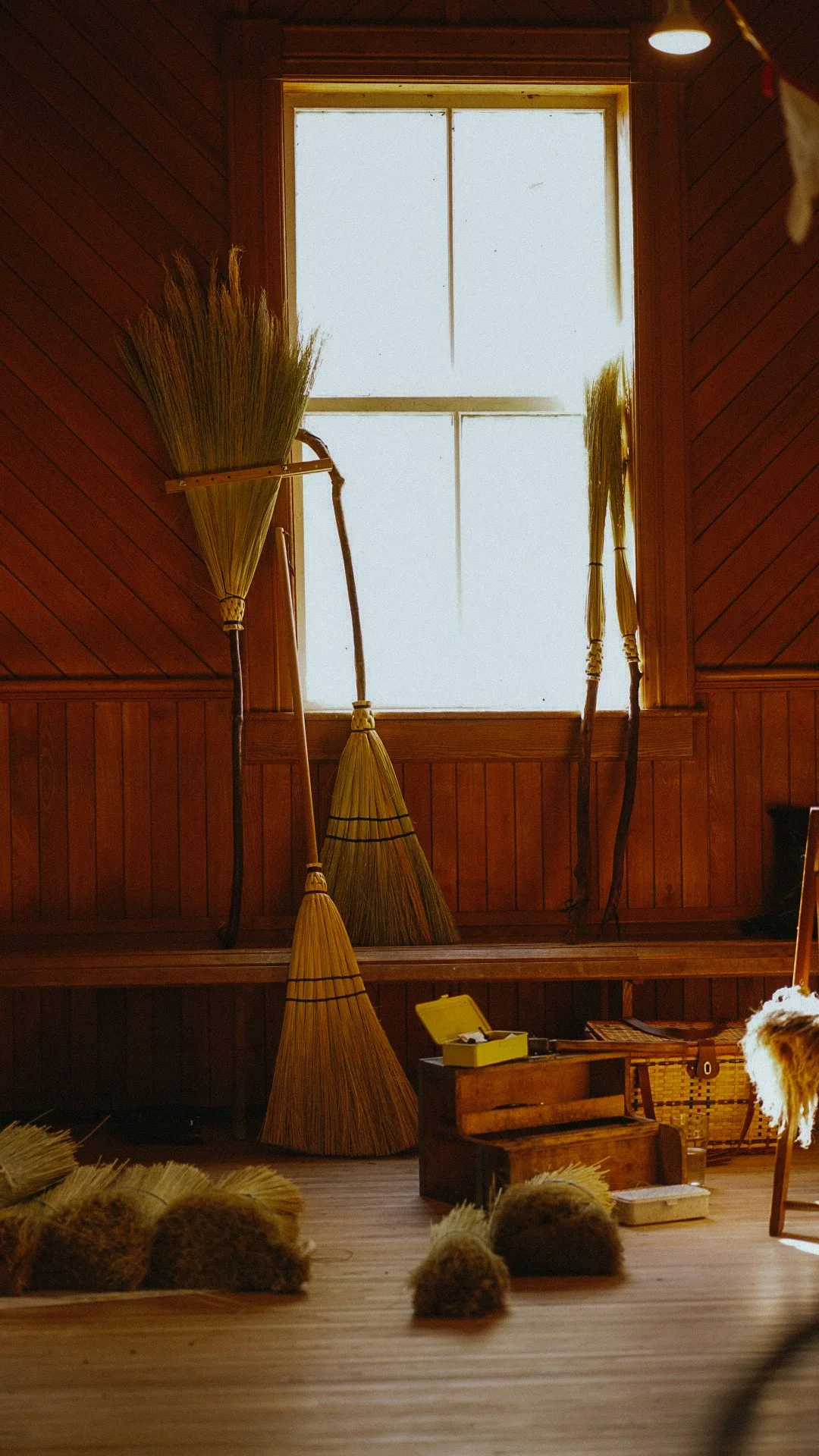 Intérieur d'une pièce en bois avec des balais en paille, une boîte à outils en bois, une caisse en osier, et un fauteuil, avec une fenêtre à carreaux en arrière-plan.