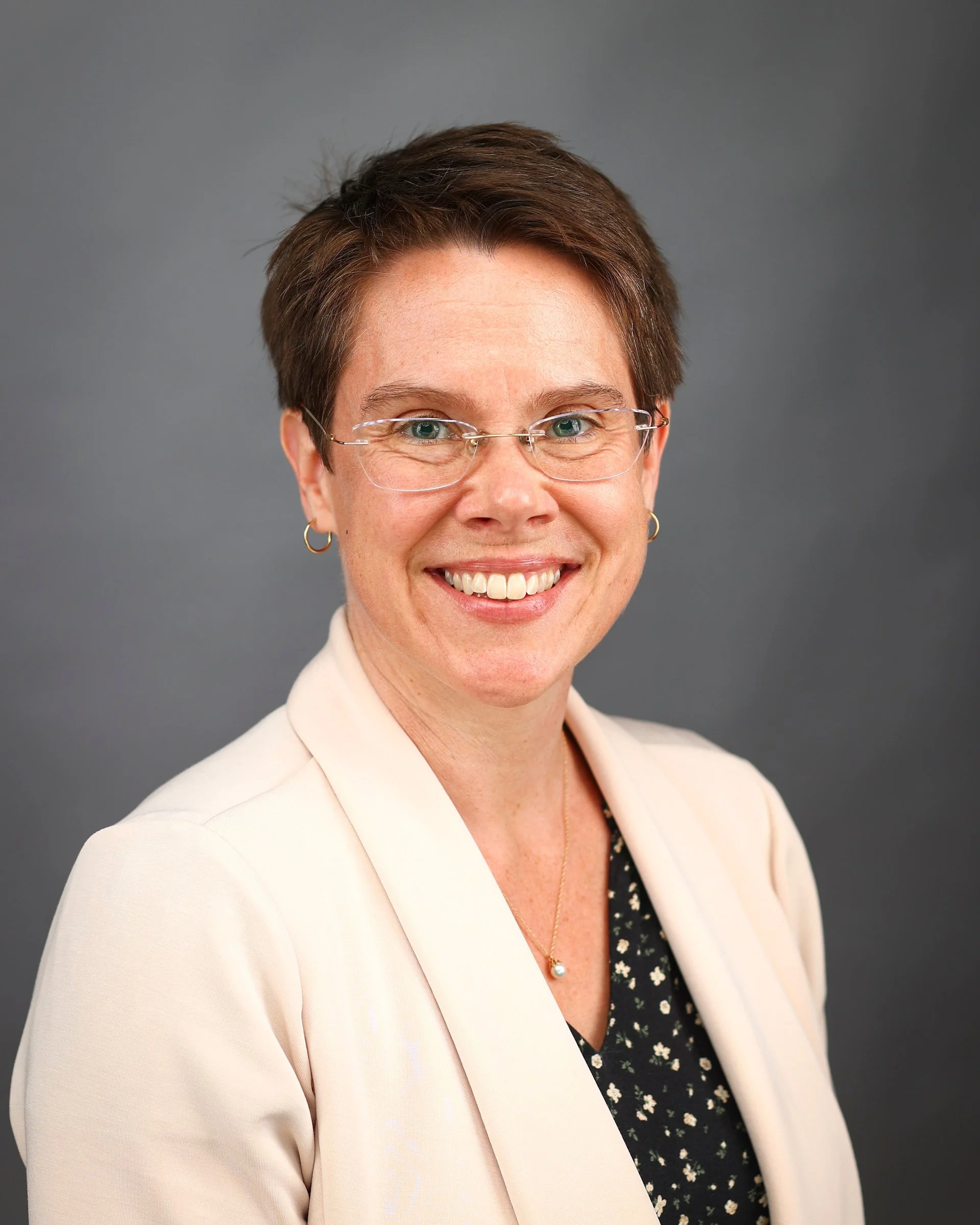 A woman with short brown hair, wearing glasses, gold hoop earrings, a pearl necklace, a beige blazer, and a black blouse with white floral pattern, smiling against a gray background.