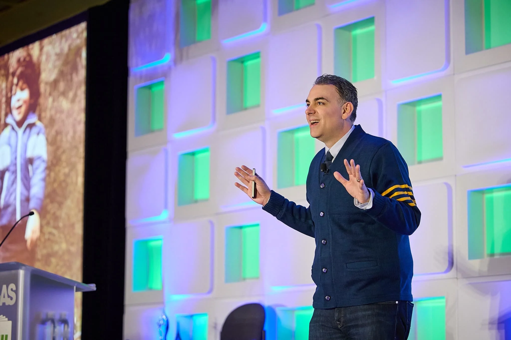 A man in a dark blue varsity-style jacket giving a presentation on a stage with colorful square patterns in the background.