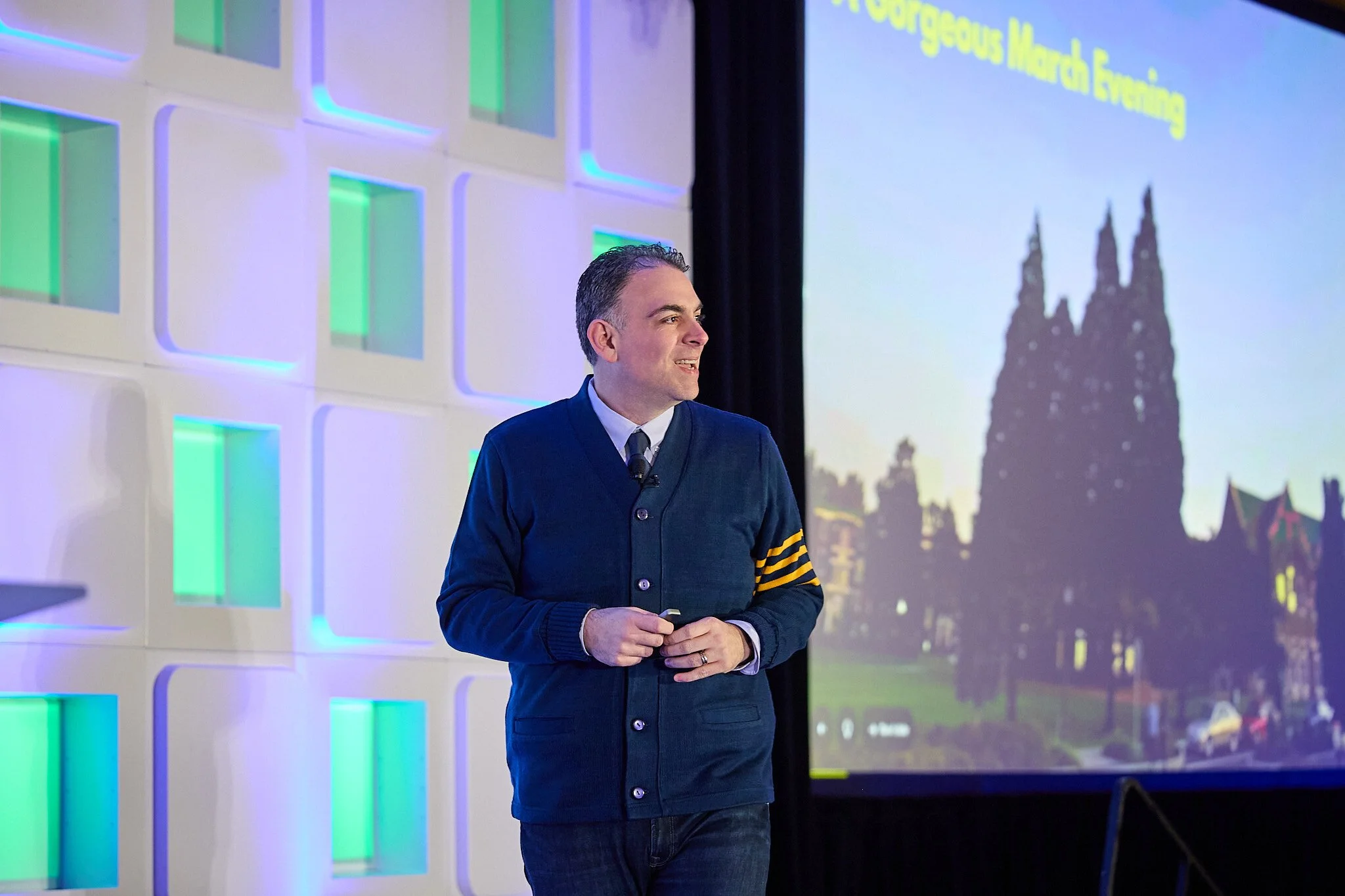 A man in a dark blue cardigan with yellow stripes on the arm, wearing a light blue shirt and dark jeans, standing in front of a large screen displaying a colorful presentation slide, smiling and holding a small device in his hands.