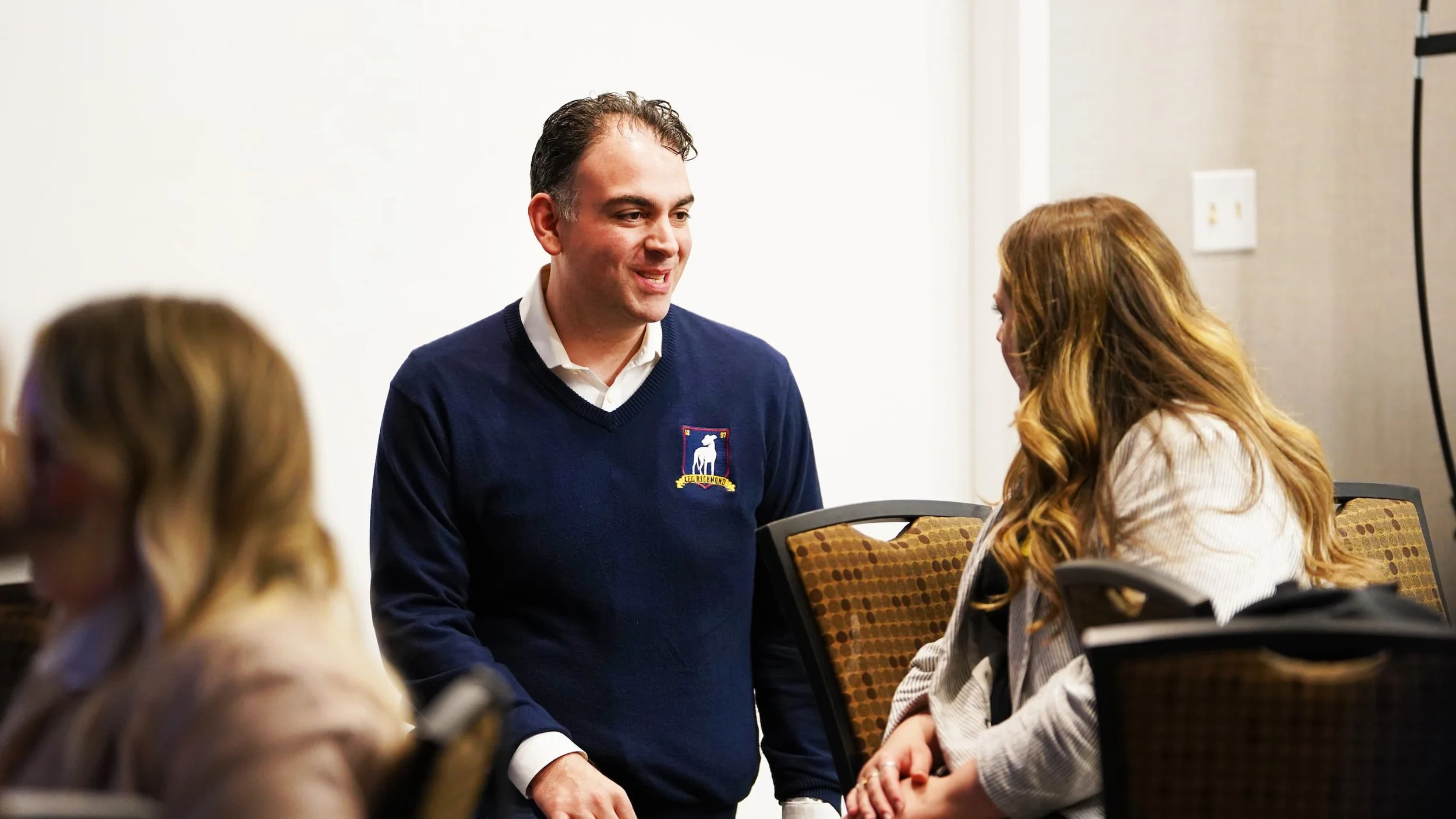 Man in a blue sweater talking to a woman at a conference table, with other women in the foreground and a white wall in the background.