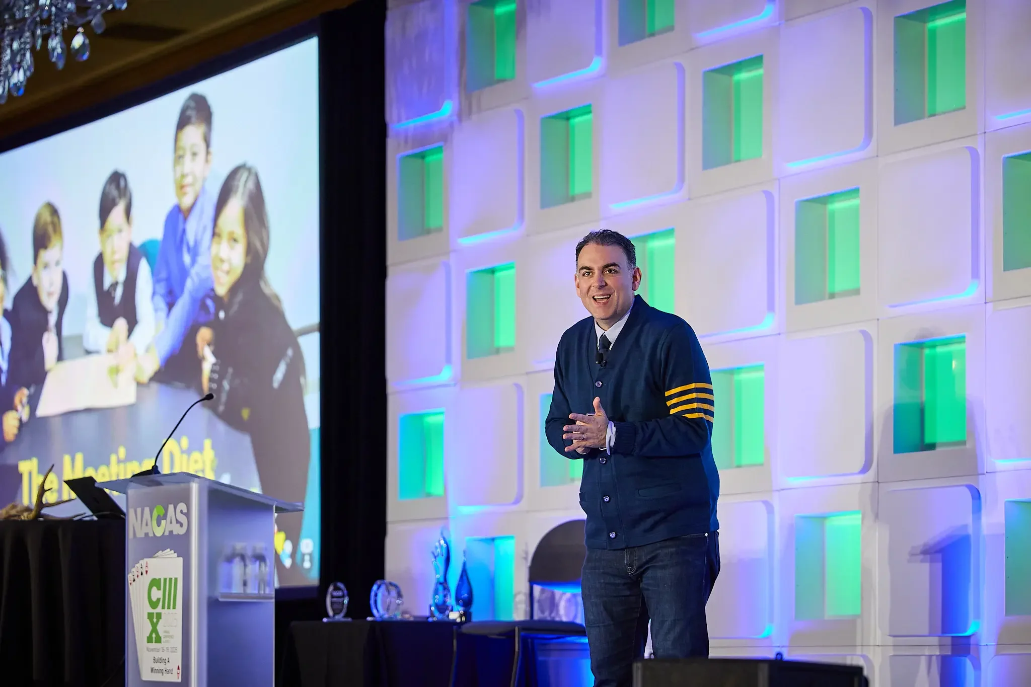 A man in a dark blazer and jeans is standing on a stage at a conference, speaking to an audience. Behind him is a large screen displaying a group photo of several children and a colorful, modern wall with green and blue accents.