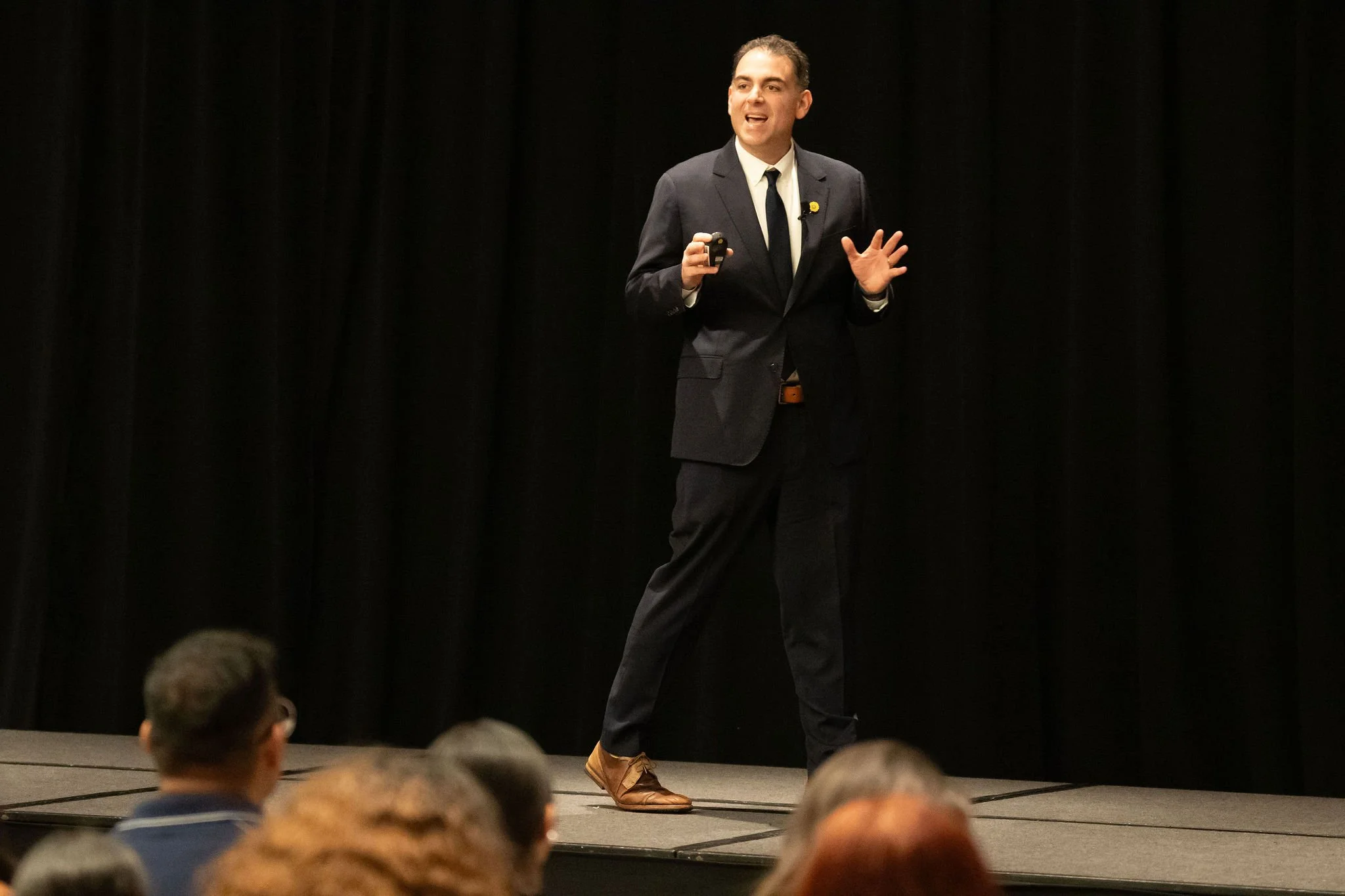 Man in dark suit and tie giving a presentation on stage in front of a black curtain, with audience members in the foreground.