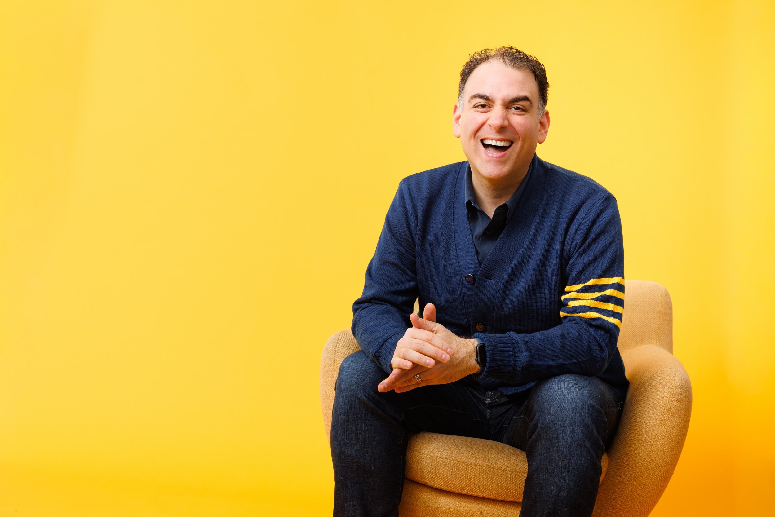 A man with short dark hair smiling and wearing a dark blue cardigan with yellow stripes on the sleeves, sitting on a yellow armchair against a bright yellow background.