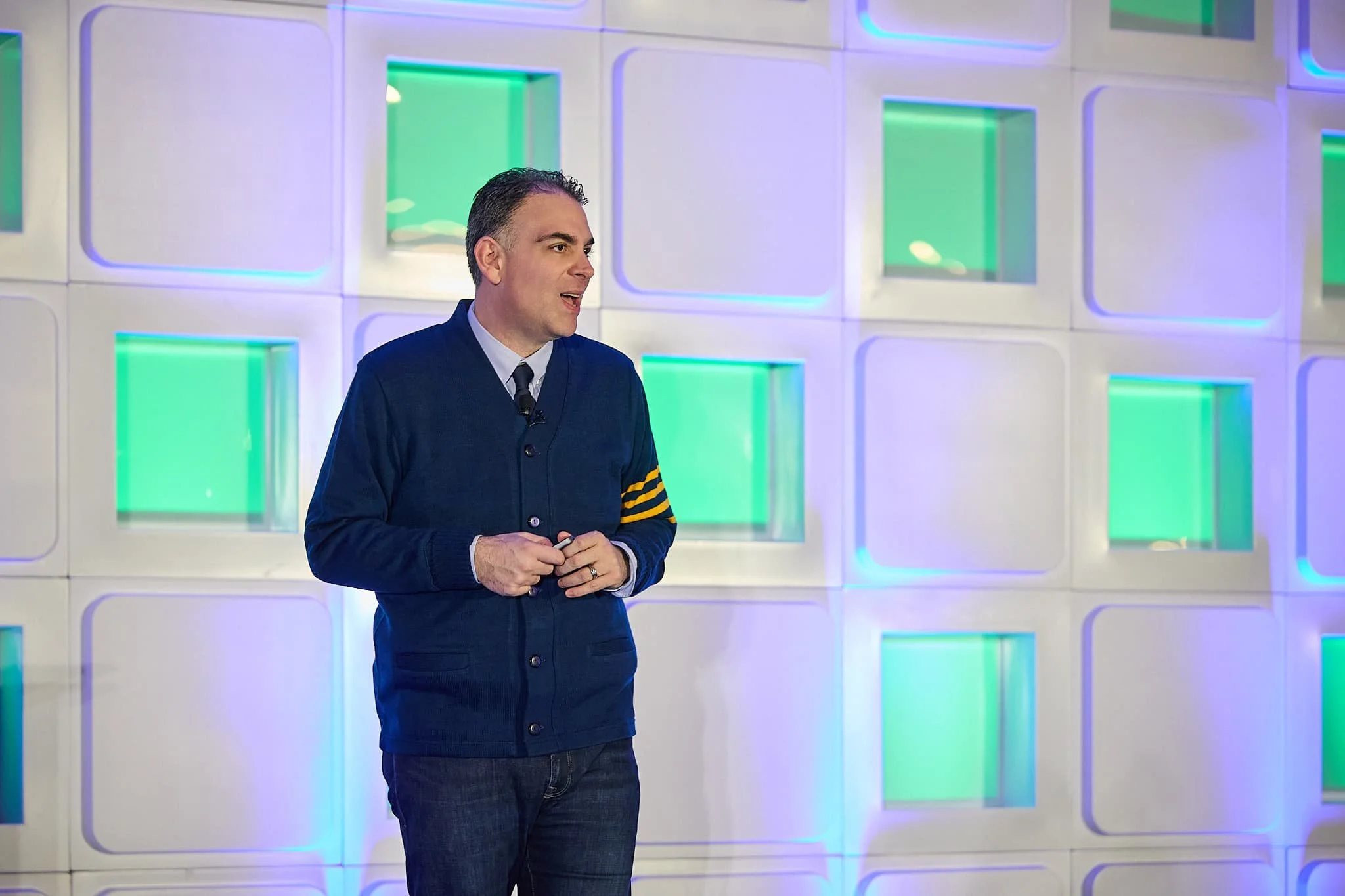 A man wearing a dark blue cardigan with yellow stripes on the sleeve stands in front of a white wall with illuminated green and blue square panels, speaking or presenting.