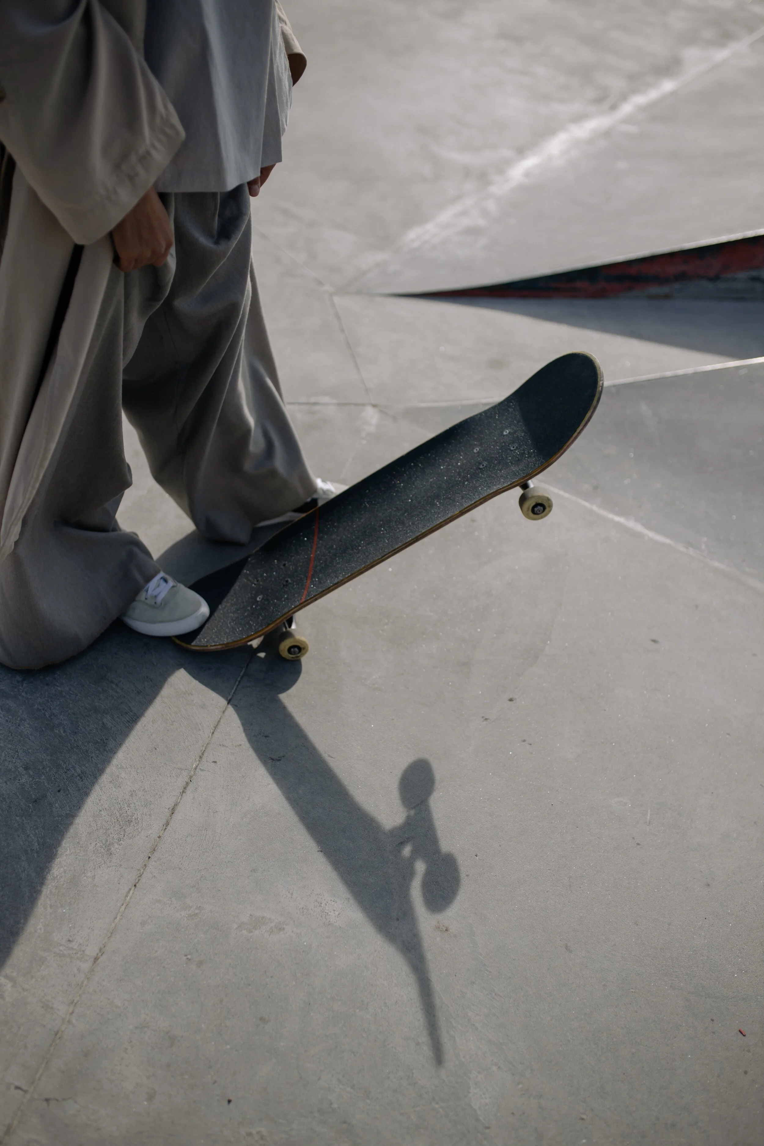 Close-up of a skateboarder’s legs and skateboard on a concrete skatepark, with long shadows cast on the ground.