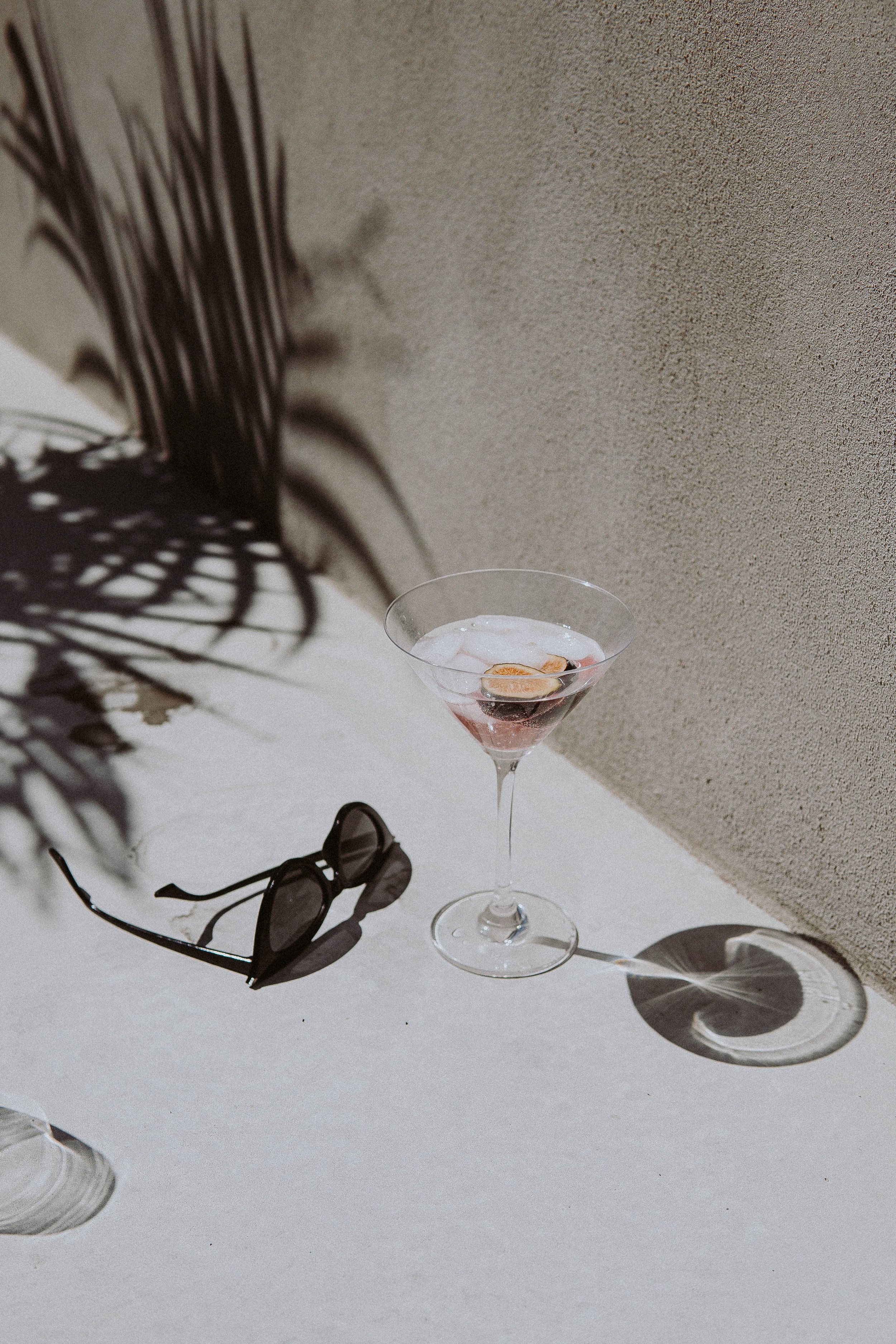 A martini glass with pink cocktail and garnish, sunglasses and shadow of leaves on a white surface, against a beige textured wall.