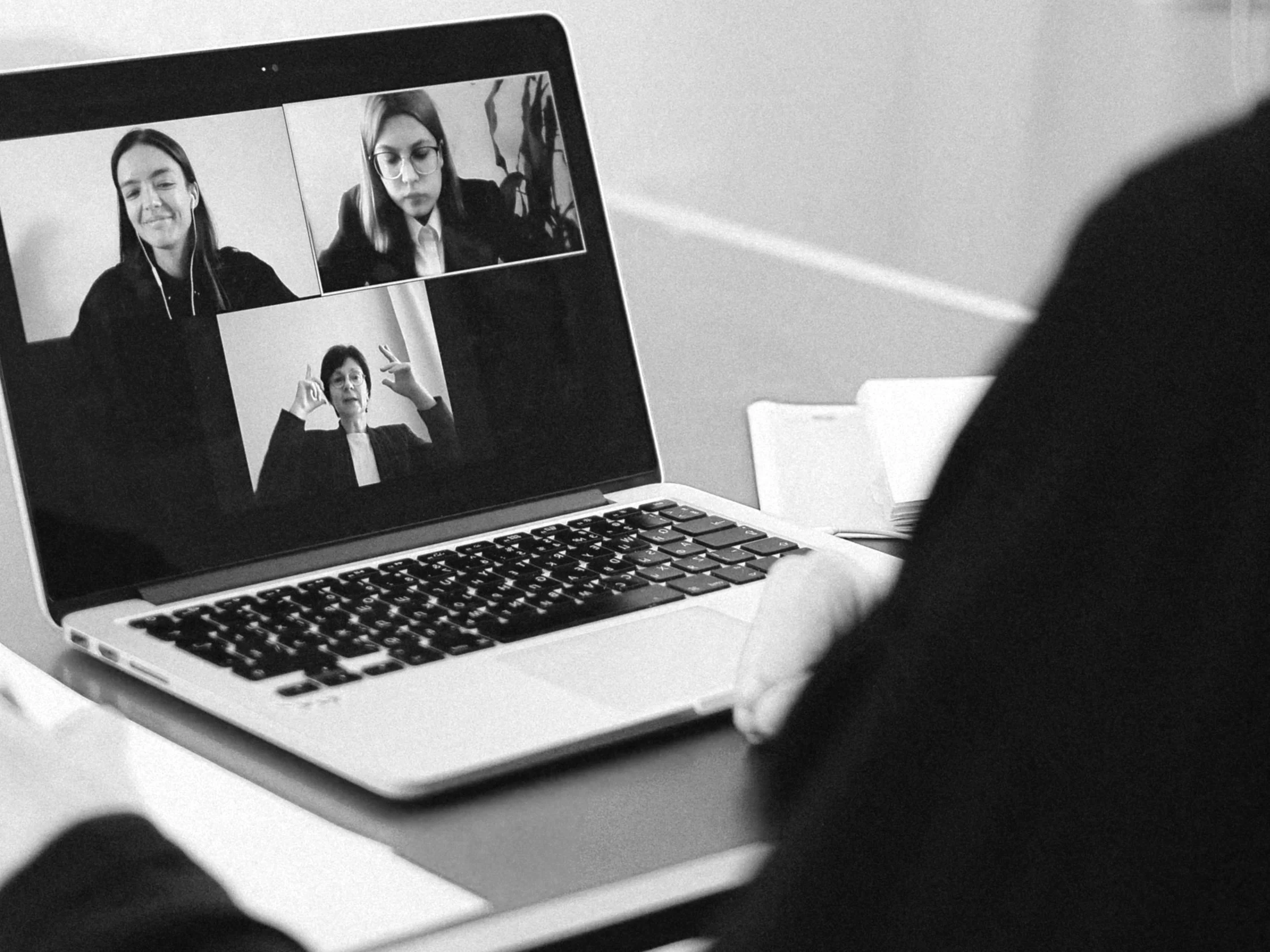A person in business attire participating in a virtual meeting on a laptop with three women on the screen, two of whom are wearing glasses and raising their hands.