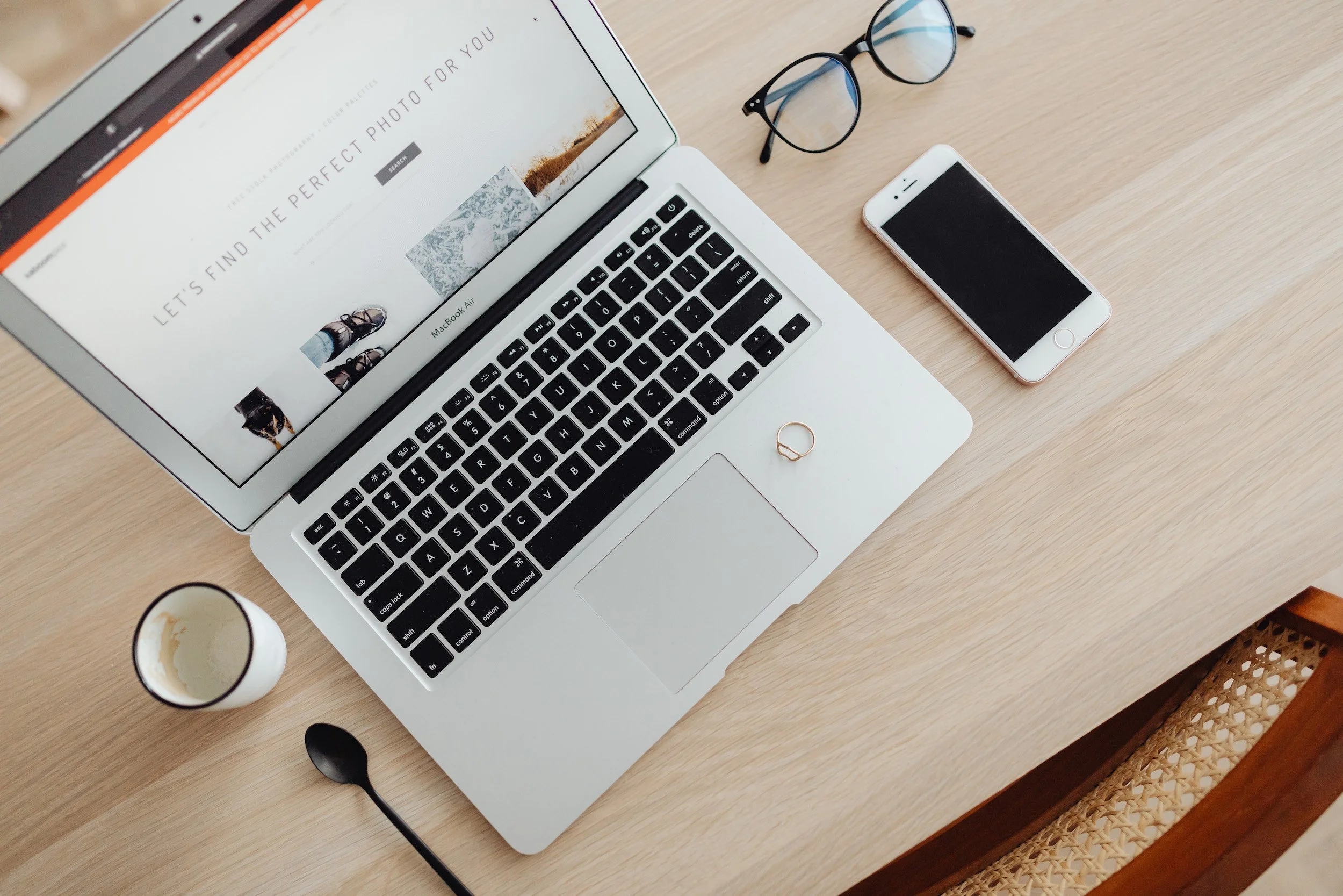 Top-down view of a wooden desk with a silver laptop displaying a webpage, a pair of black-rimmed glasses, a white smartphone with black screen, a white mug with some coffee, and a black spoon.