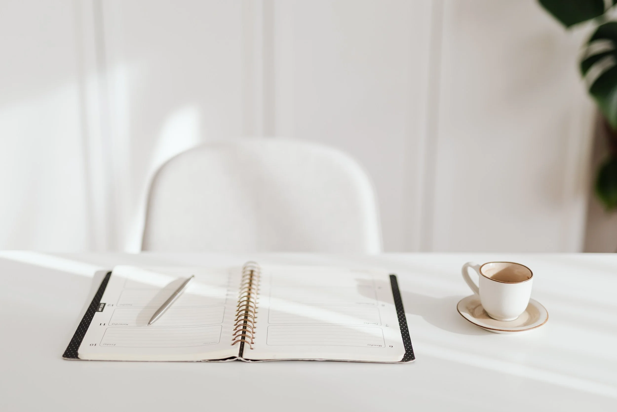 Open planner with a silver pen on a white table, and a white coffee cup with a saucer beside it, in a bright room with a white chair and a green plant in the background.