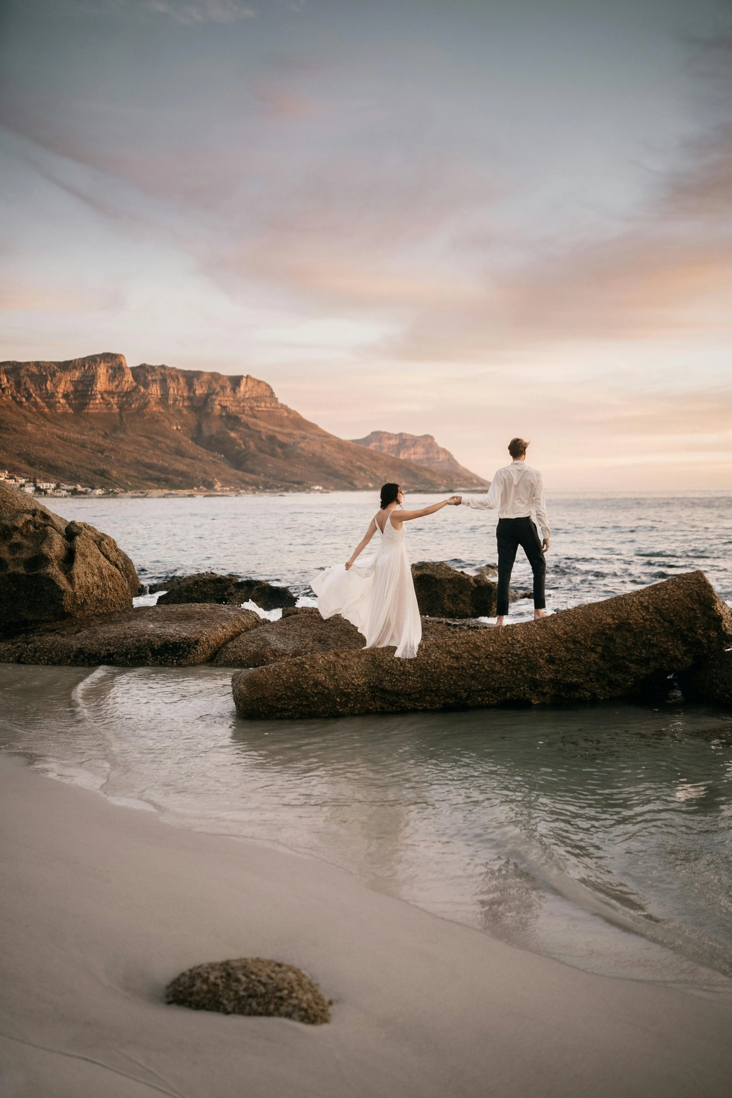A bride in a white wedding dress and a groom in a white shirt and dark pants holding hands on rocky beach at sunset, with mountains and ocean in the background.