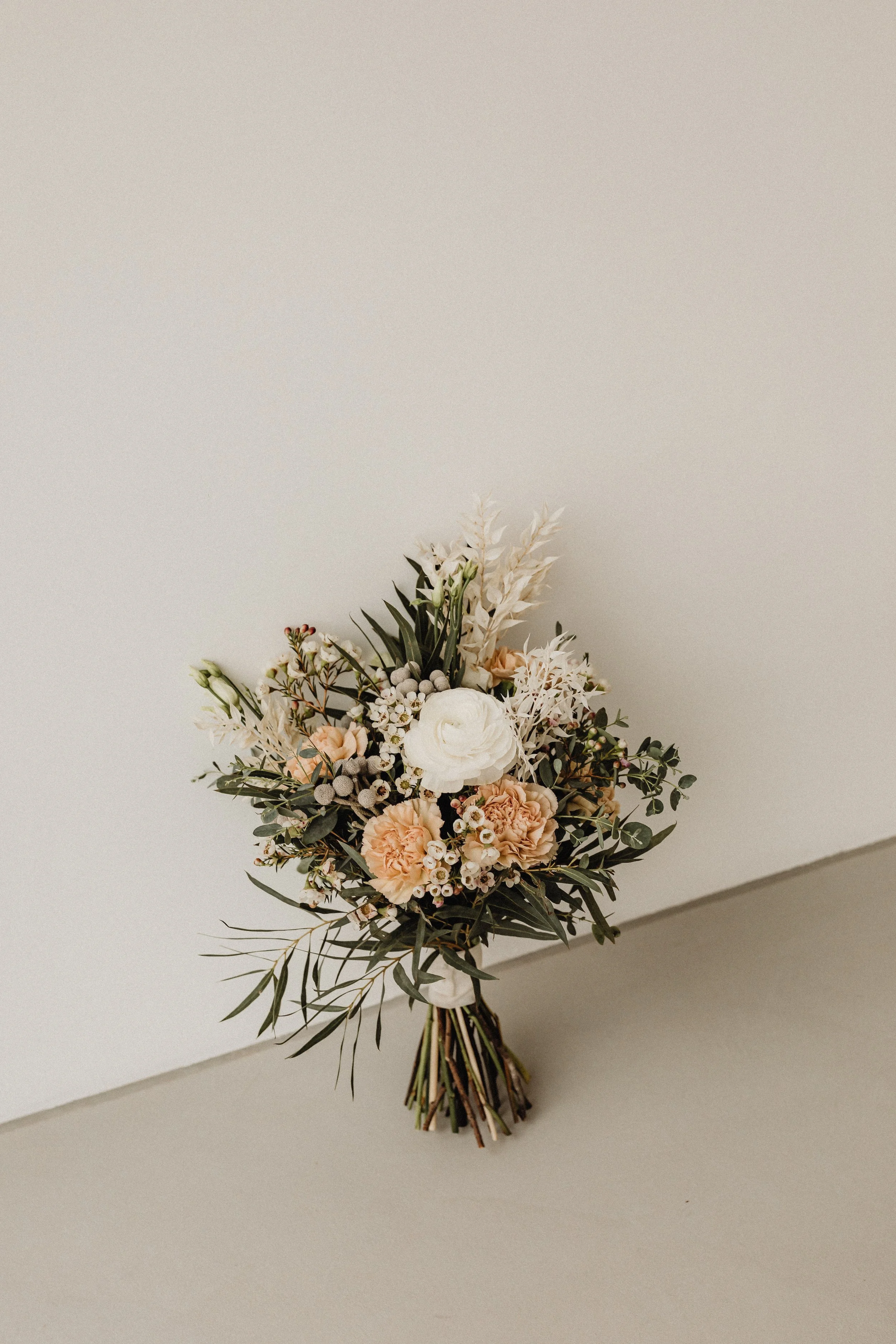 A bouquet of flowers including white, peach, and cream-colored blooms, accented with greenery, resting on a neutral-colored surface against a plain wall.