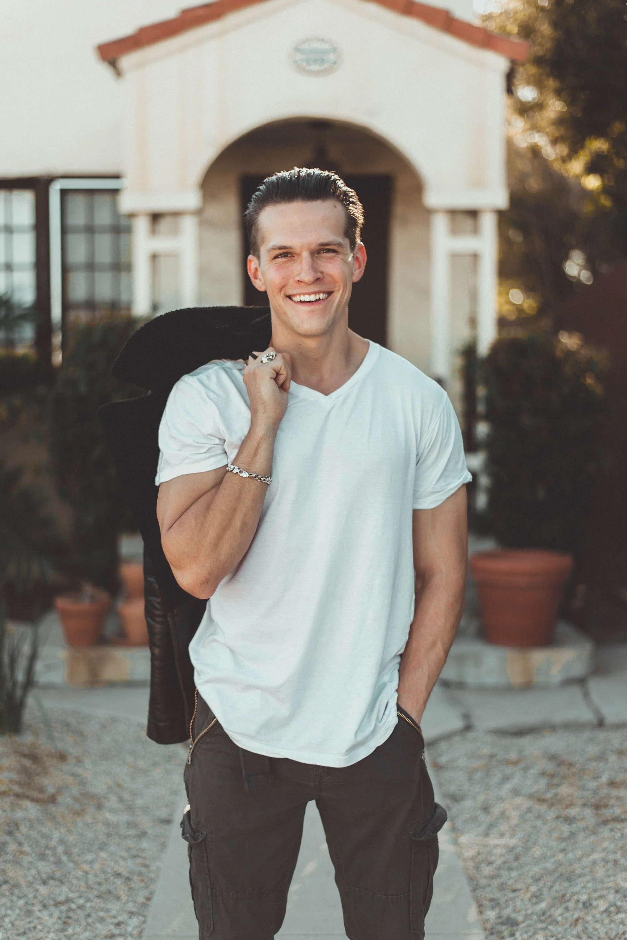 A young man with dark hair, wearing a white t-shirt, smiling while holding a black leather jacket over his shoulder, standing outdoors in front of a building with potted plants and a pathway.