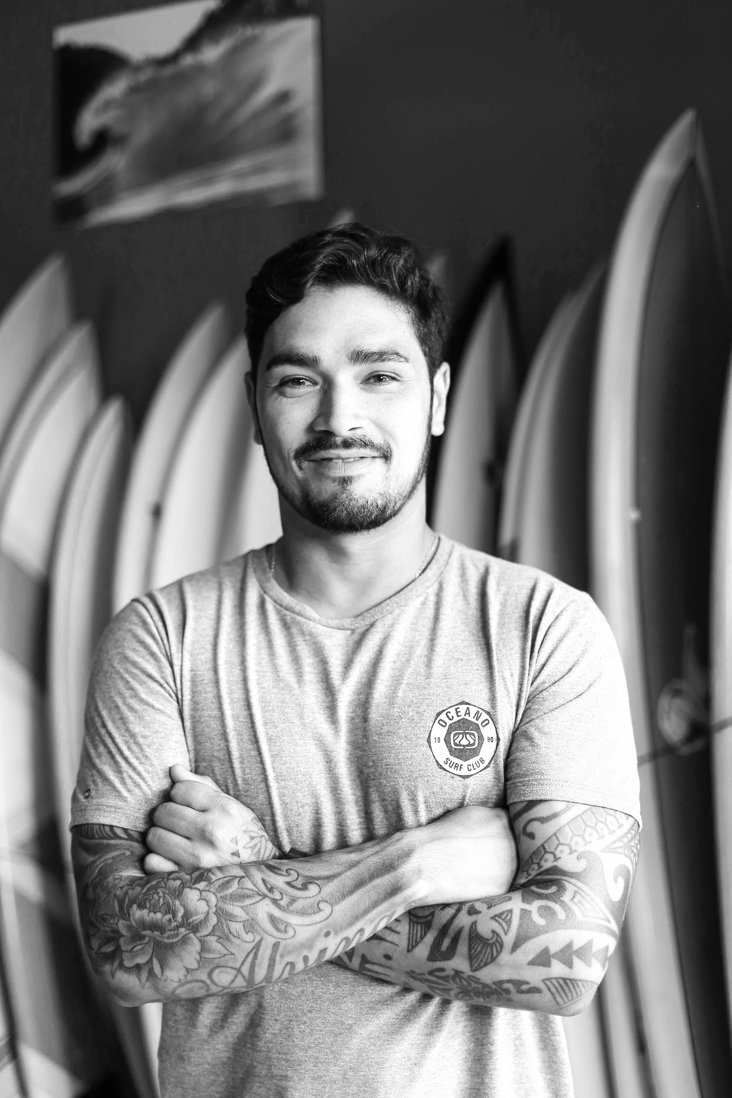 A smiling young man with tattoos on his arms, crossing his arms, standing in front of surfboards and a wall with a painting of an eagle in a surf shop.