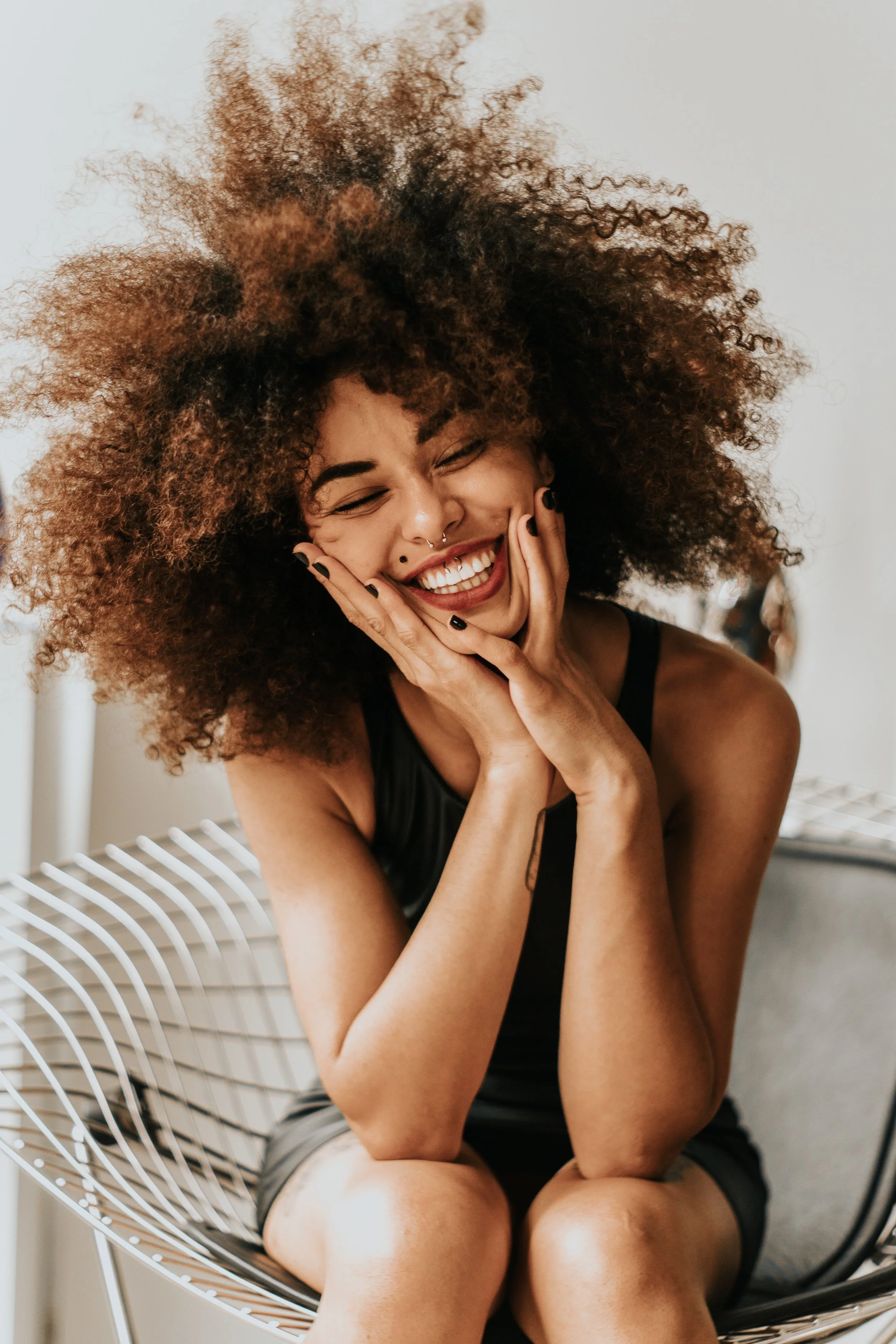 A woman with curly hair laughing and smiling with her hands on her cheeks, sitting on a modern wire chair.