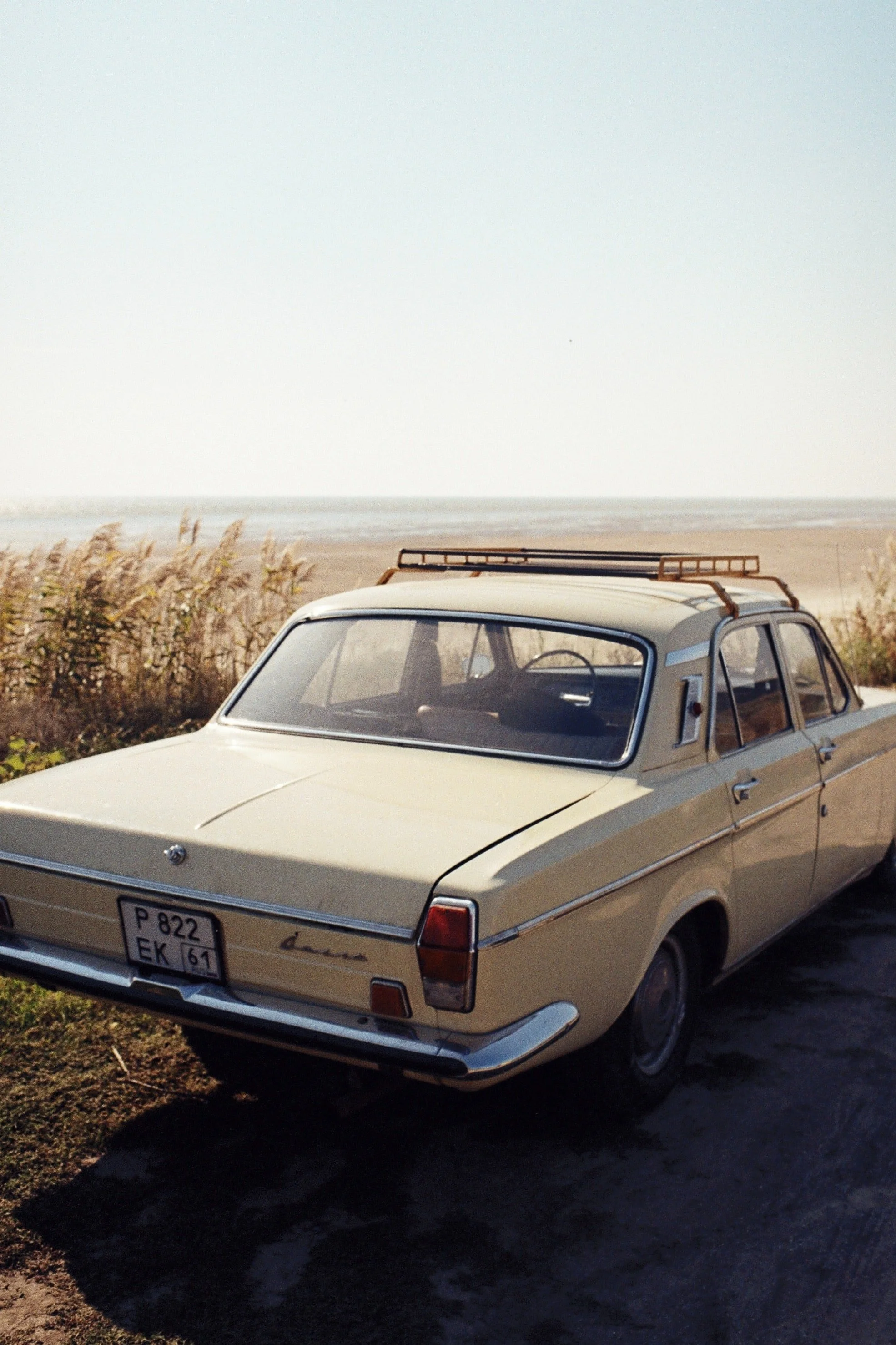 A vintage beige car parked on a dirt area near a beach with grass and the ocean in the background.