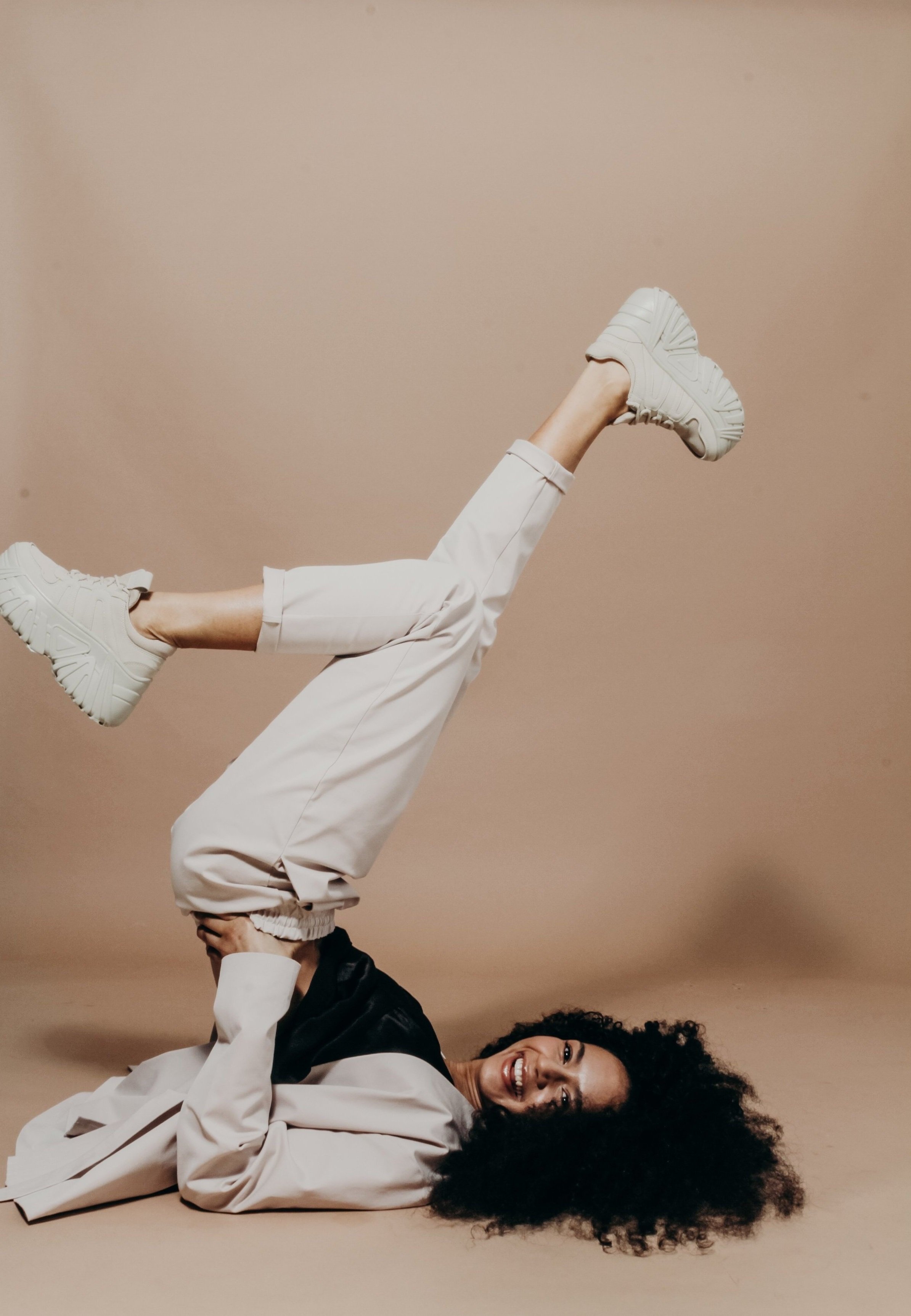 Woman lying on the ground with her legs raised and smiling while wearing white sneakers, beige pants, and a beige and black jacket against a beige background.