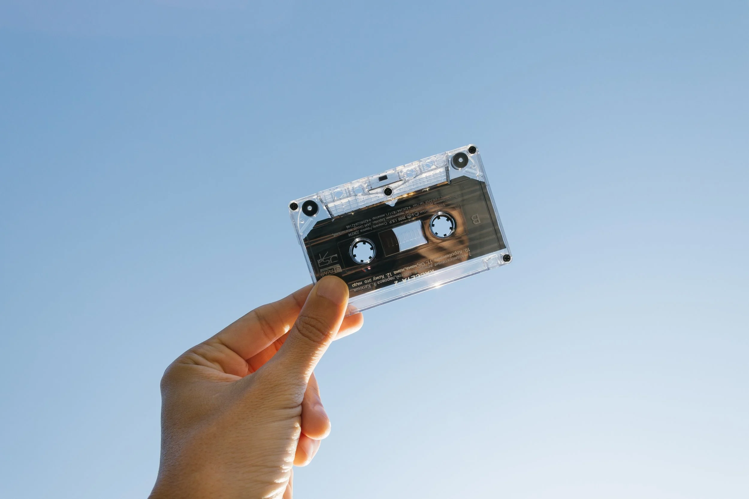 Hand holding a clear audio cassette tape against a blue sky background.