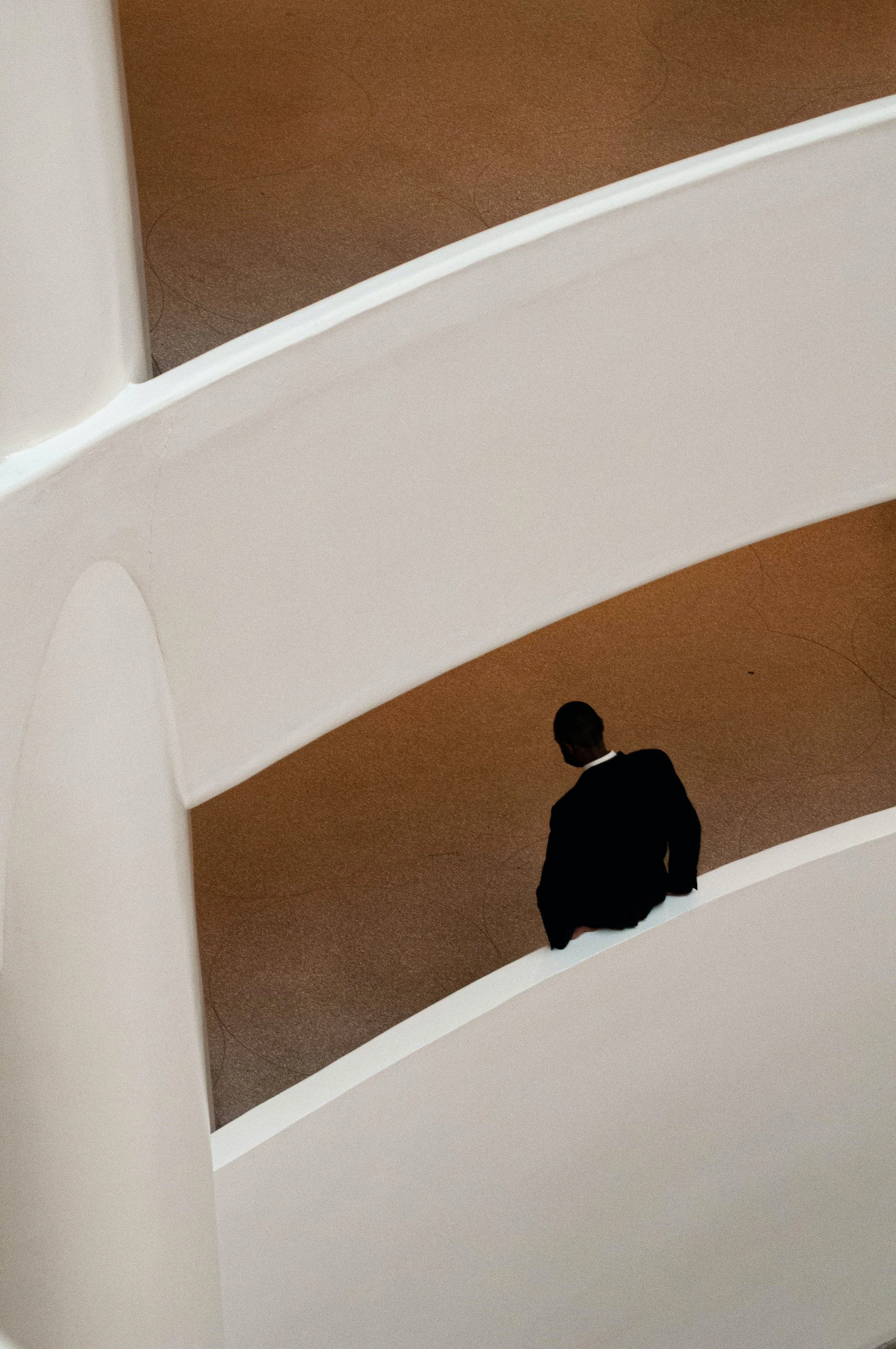 A man in a dark suit standing on a modern, curved white balcony in a building with beige walls.