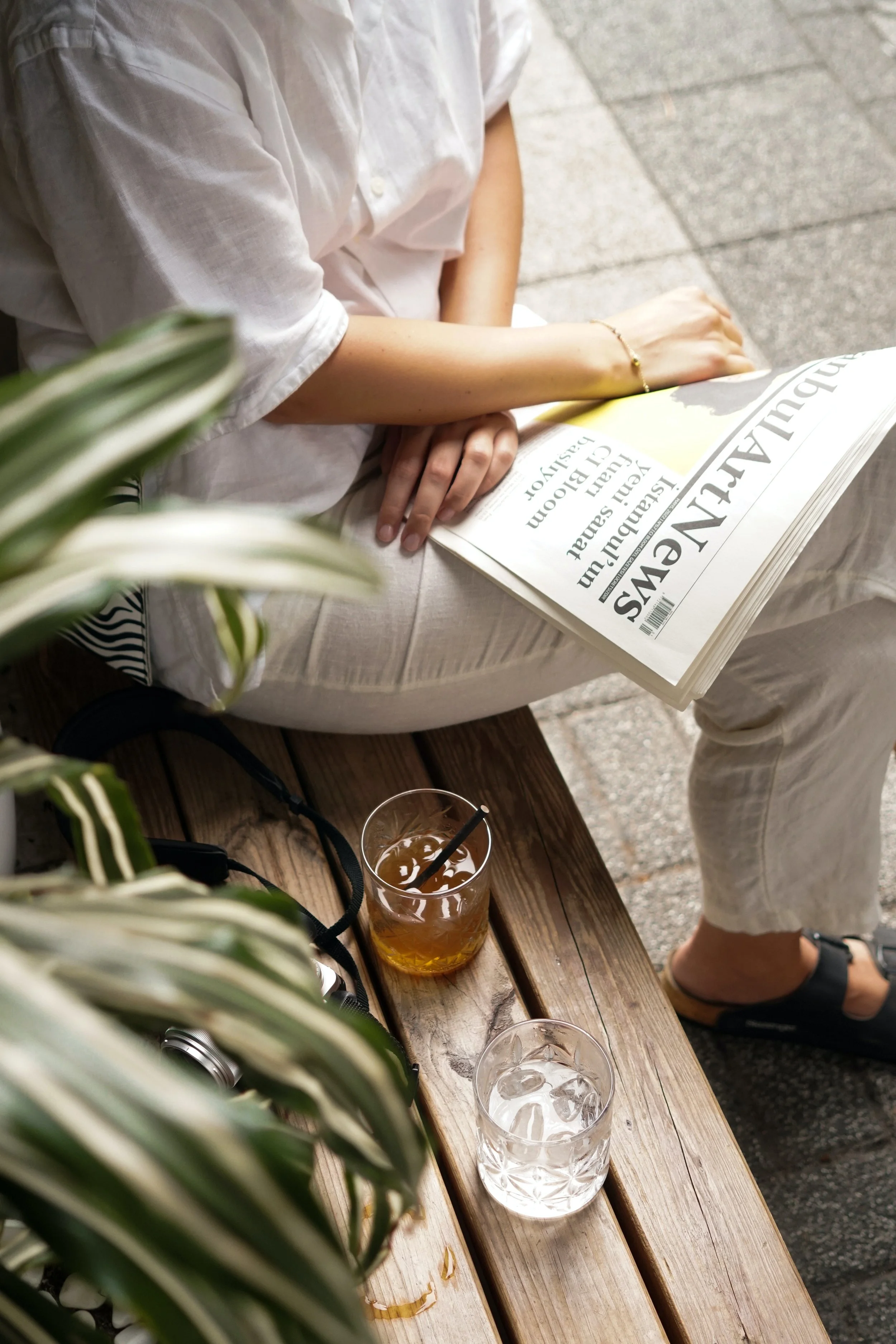 Person sitting on a wooden bench, reading a newspaper with glasses of water and iced tea on the table.