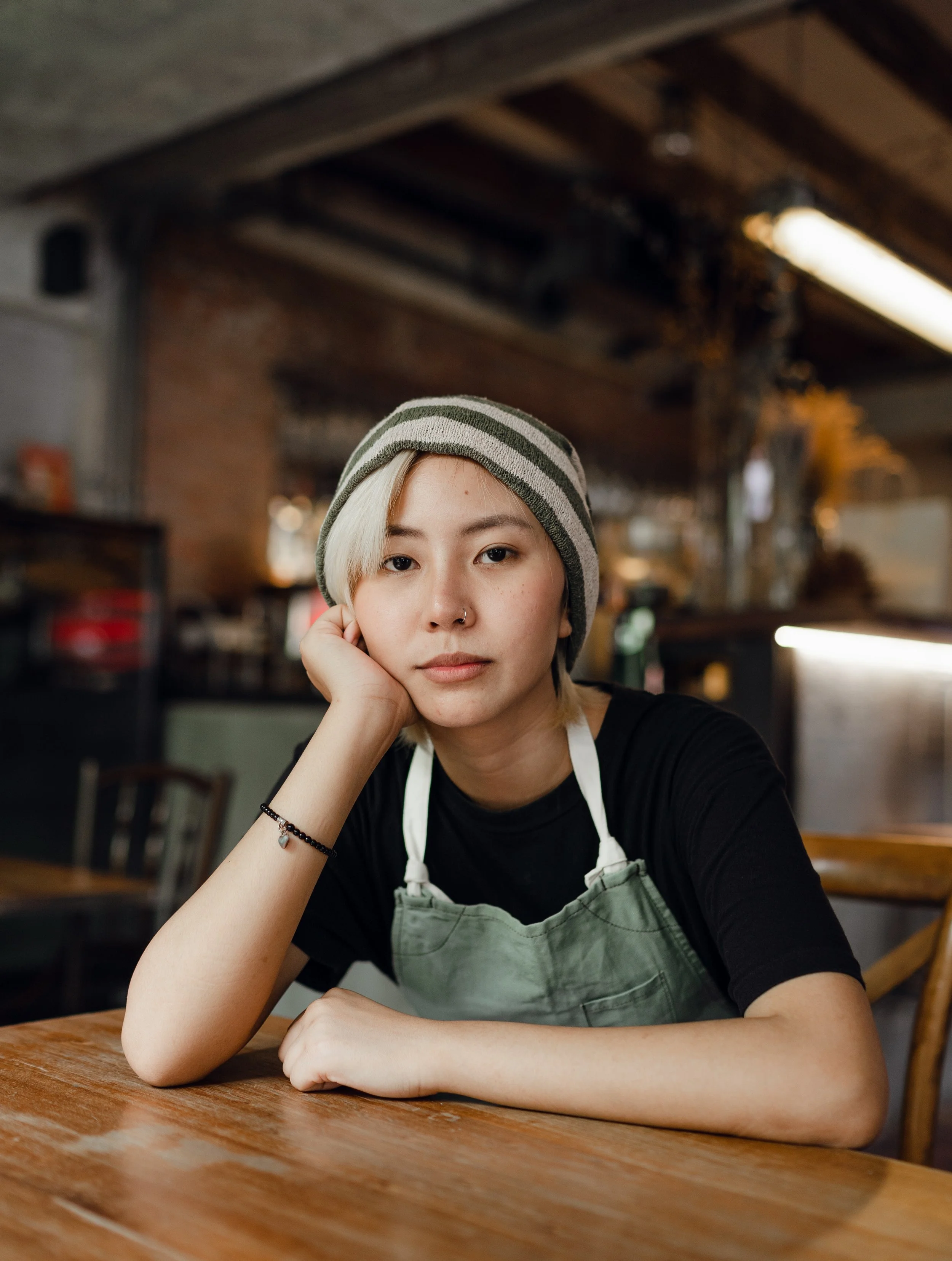 A young woman with short blonde hair and a nose piercing, sitting at a wooden table in a cozy, rustic restaurant. She is wearing a striped beanie, a black shirt, and a green apron, resting her chin on her hand and looking at the camera.