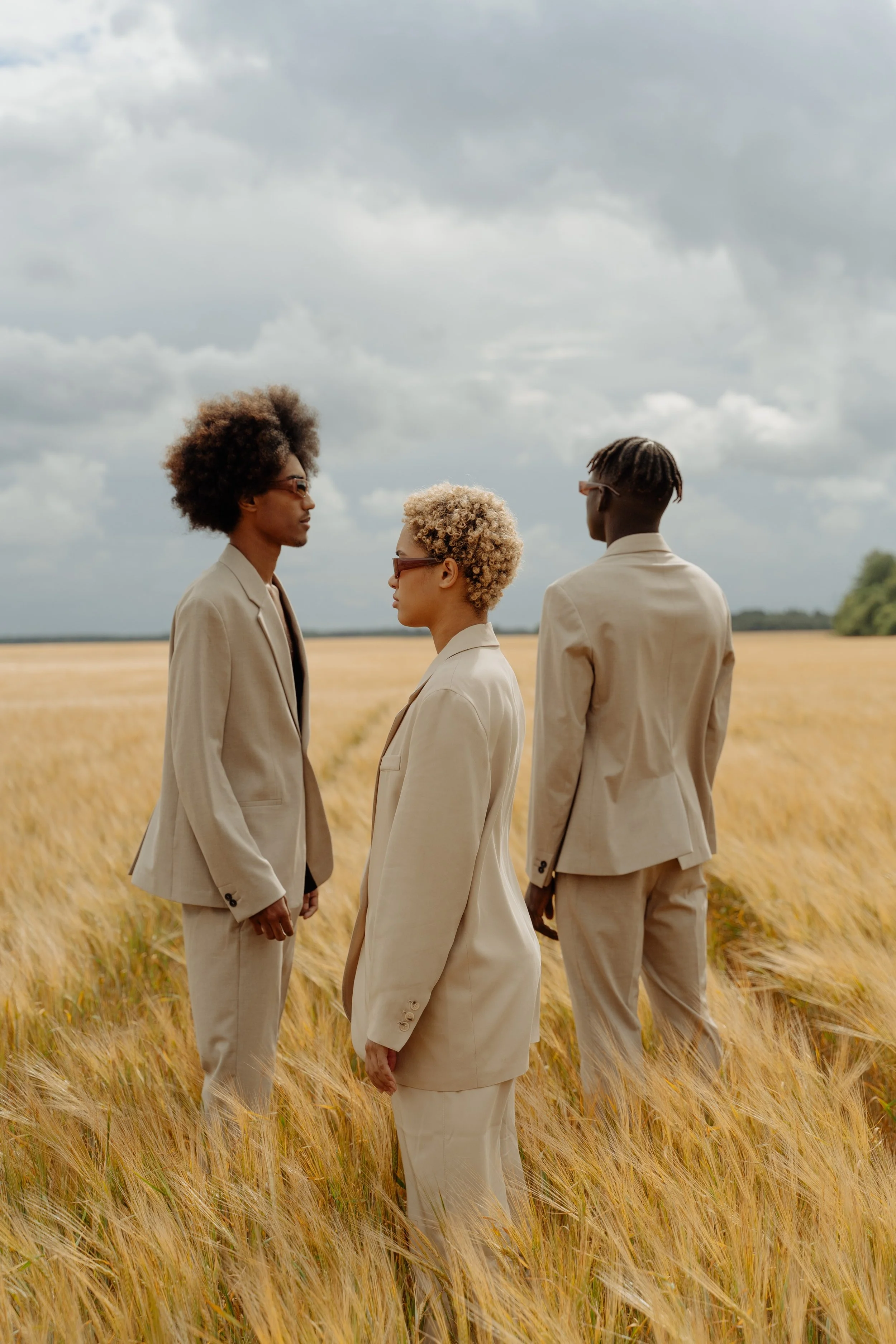 Three people in beige suits standing in a golden wheat field under a cloudy sky.
