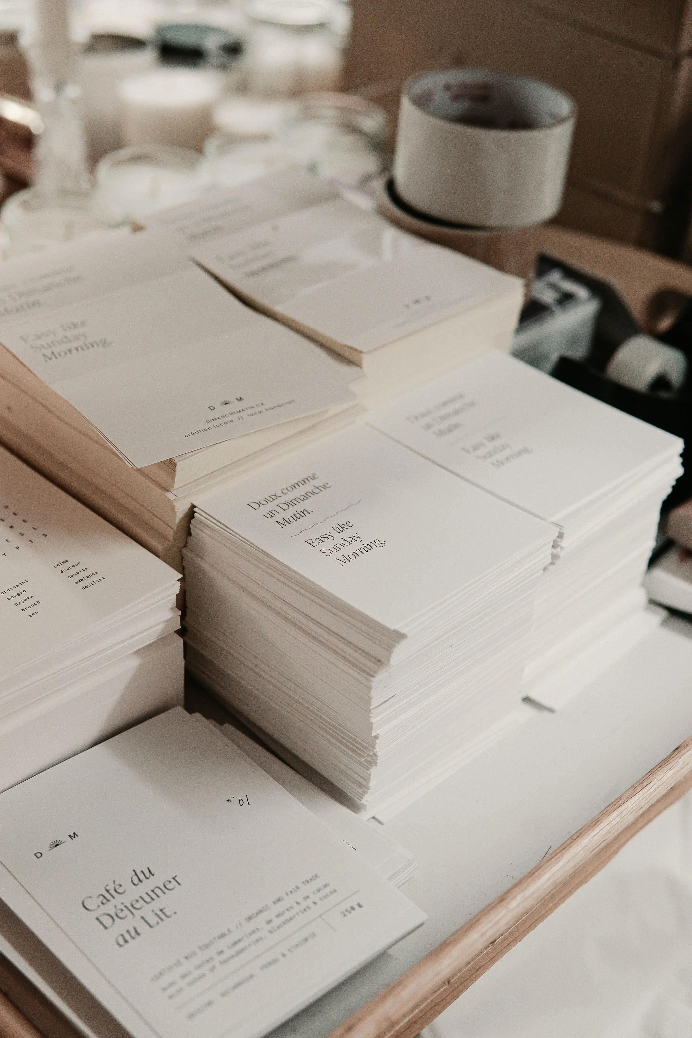 Stacks of white paper cards with printed text, on a wooden tray, with rolls of tape and blurred jars in the background.