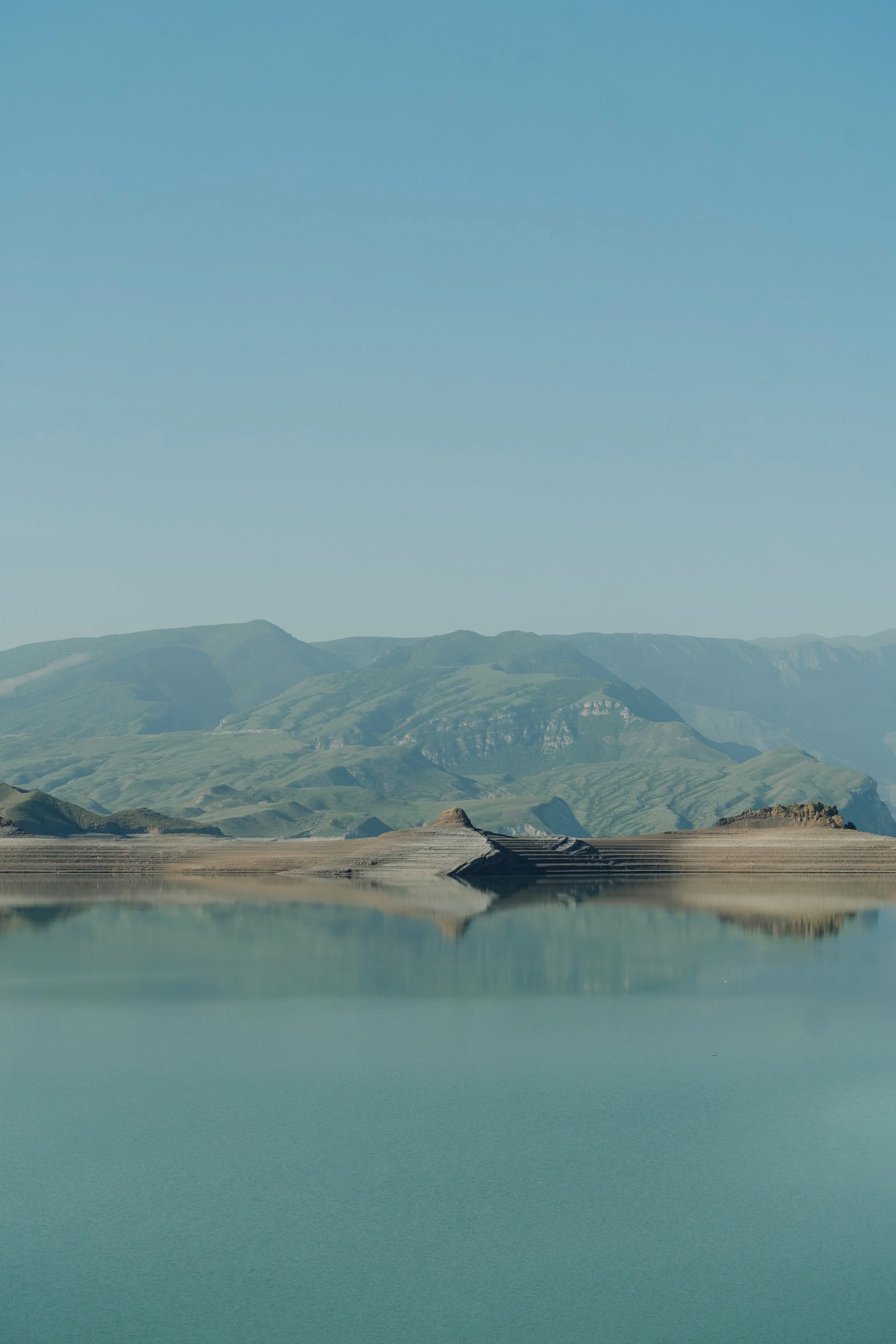 Landscape with green mountains and reflected water under a clear blue sky.