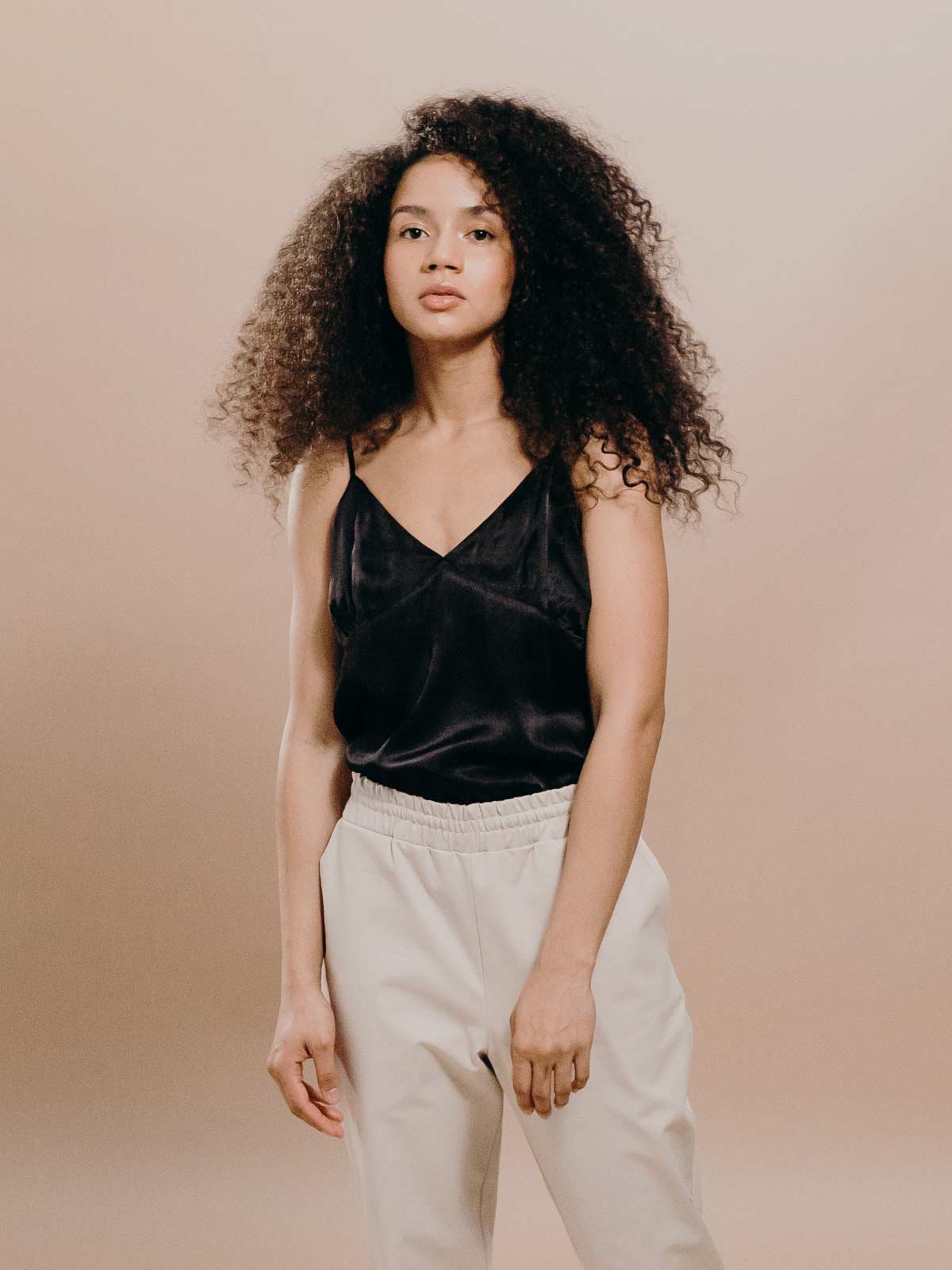A young woman with curly hair wearing a black satin spaghetti strap top and beige pants standing against a plain neutral background.