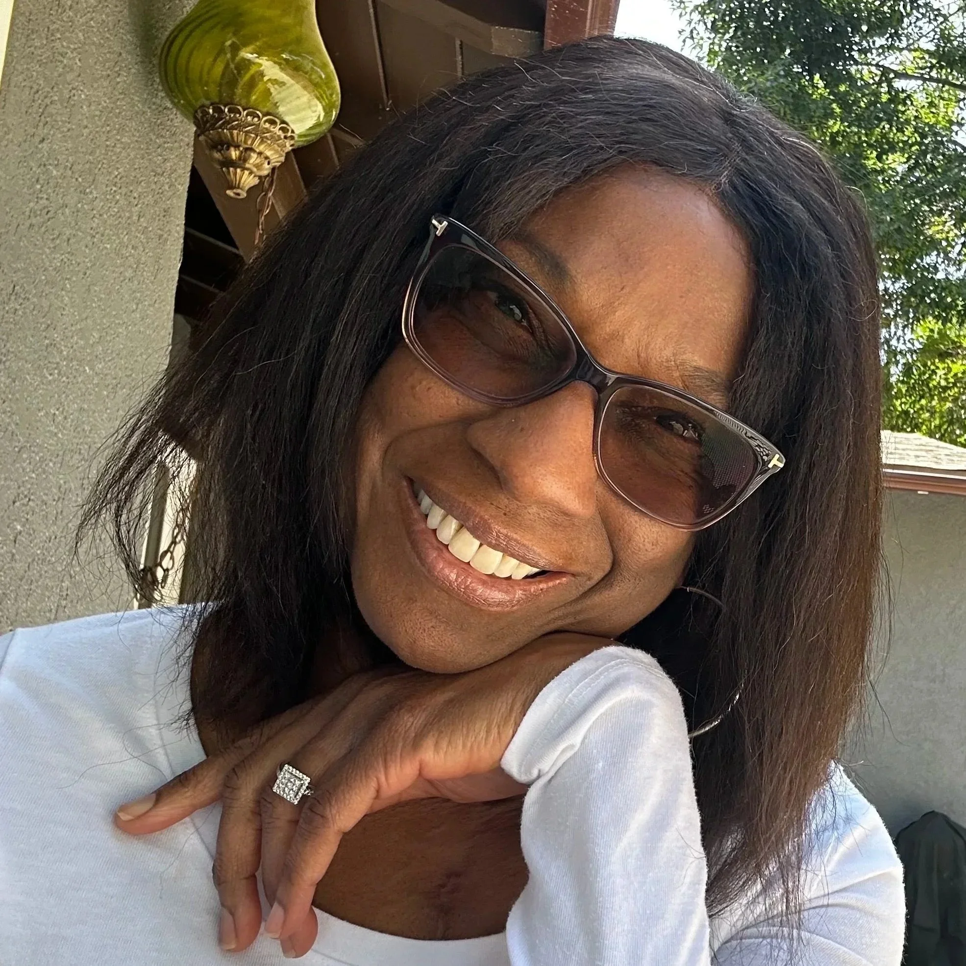 Close-up of a smiling woman with sunglasses, resting her chin on her hand, wearing a white top and a ring, outdoors in sunlight with green trees in the background.