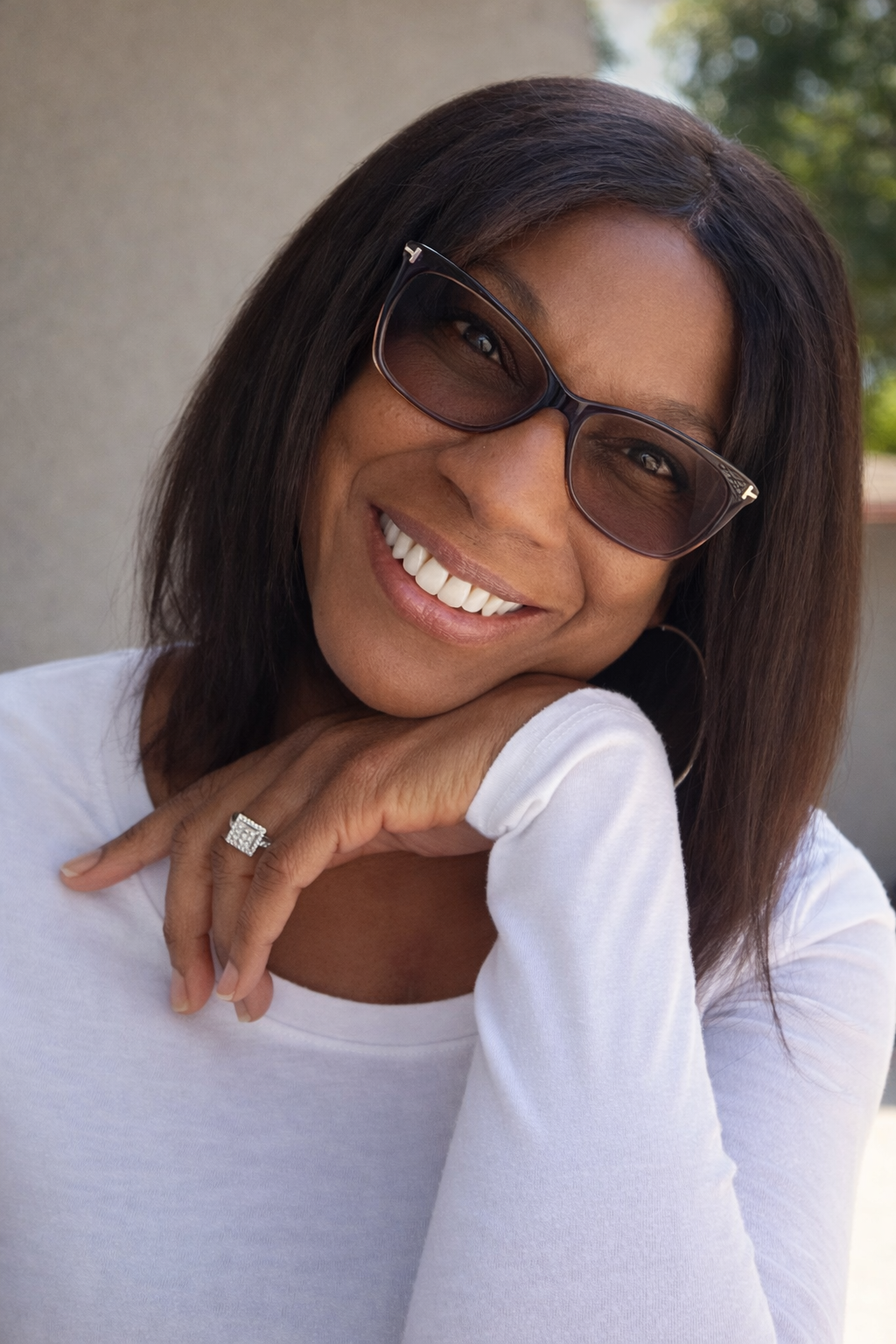 A woman with dark brown hair wearing sunglasses, a white shirt, and a diamond ring, smiling and resting her chin on her hand.