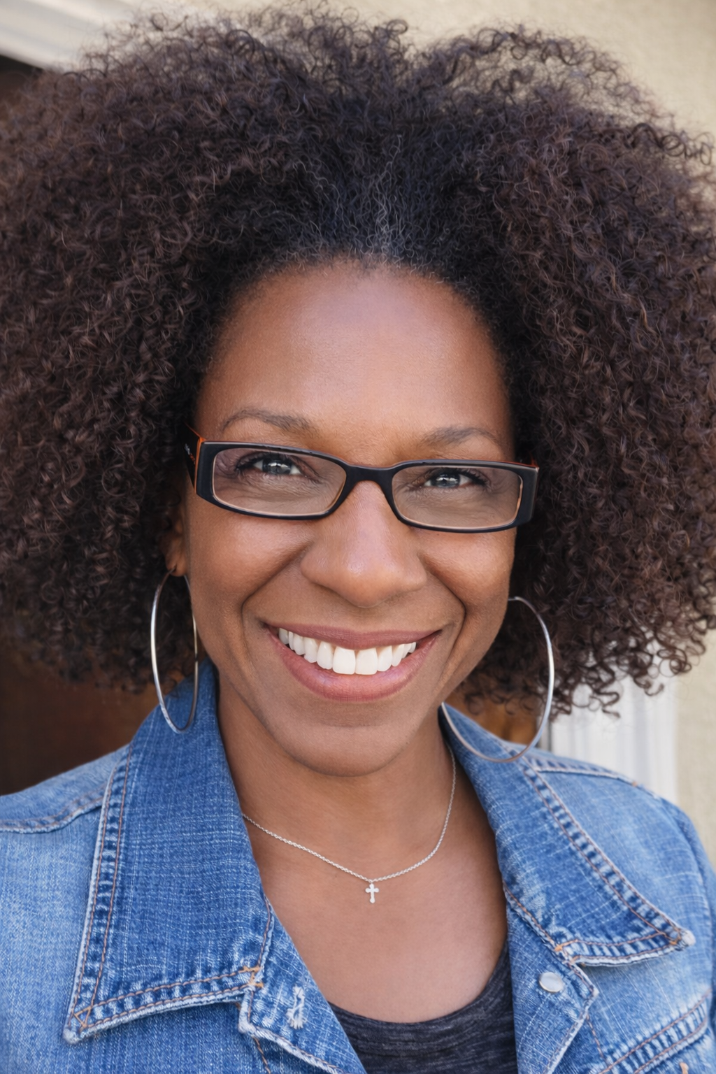 Smiling woman with curly hair, glasses, hoop earrings, denim jacket, and cross necklace.