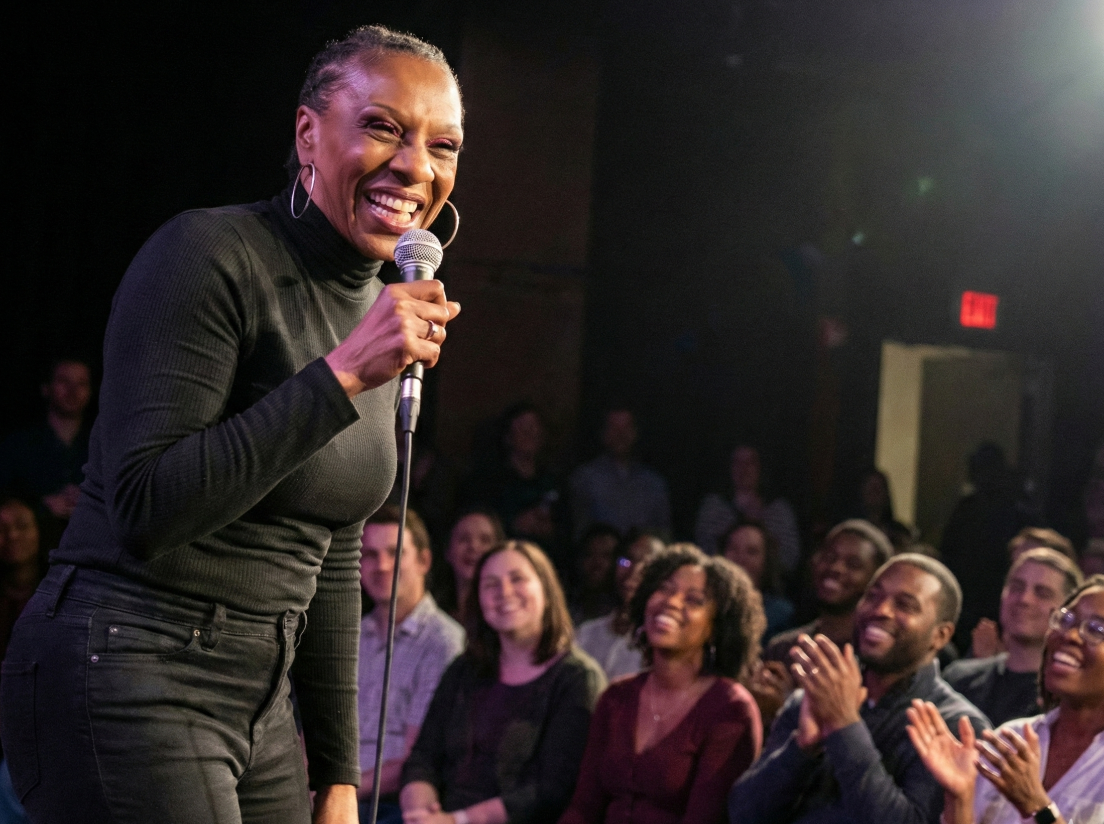 A woman laughing and holding a microphone on stage with an audience clapping and smiling in the background.