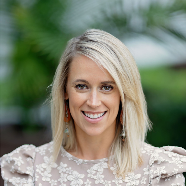 A smiling woman with blonde hair wearing a floral top outdoors with greenery in the background.