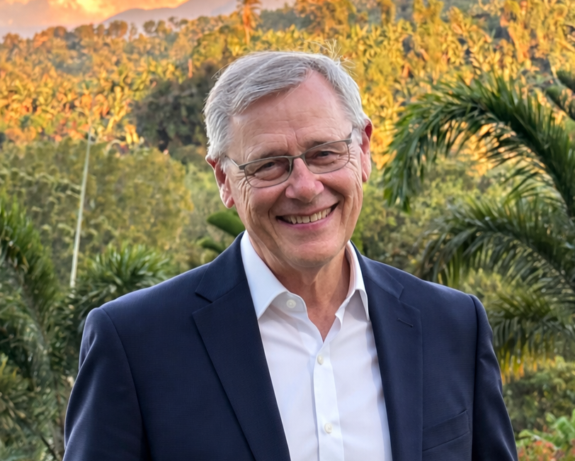 A smiling elderly man with gray hair and glasses, wearing a navy blazer and white shirt, standing outdoors with lush green trees and hills in the background during sunset.