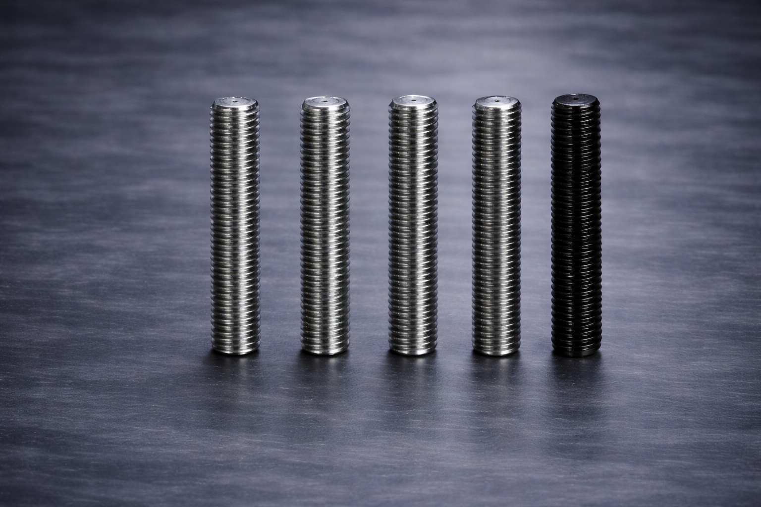 Five metal screws standing upright on a dark textured surface, with the screws gradually changing from lighter to darker shades from left to right.