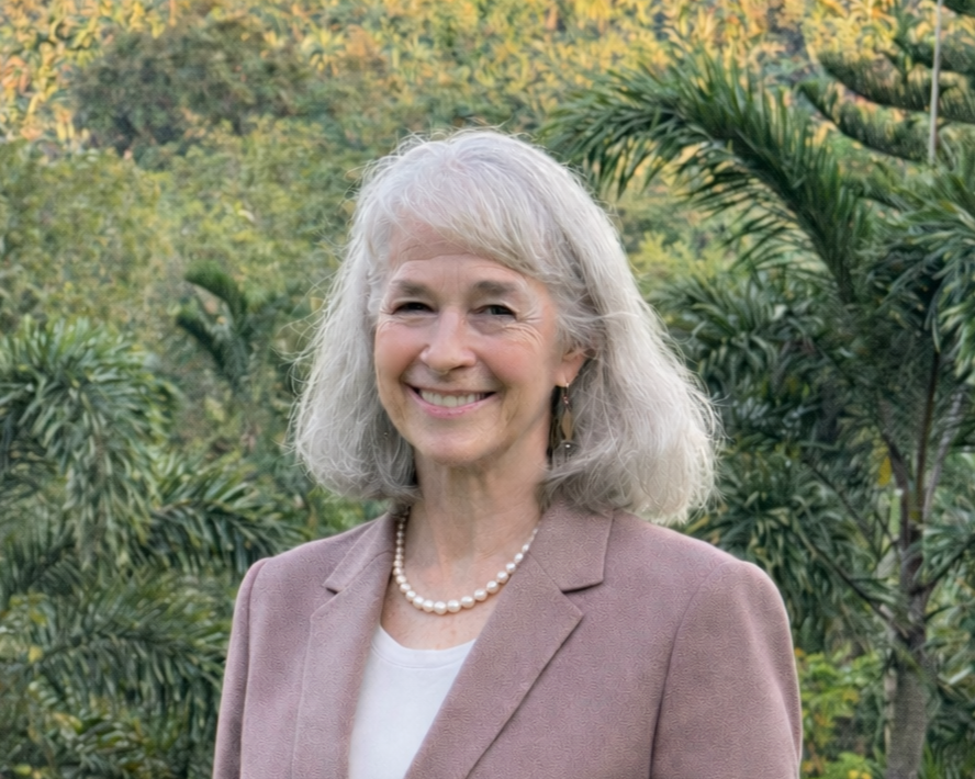 A woman with gray hair smiling outdoors, wearing a light pink blazer, white top, pearl necklace, and earrings, with green foliage in the background.