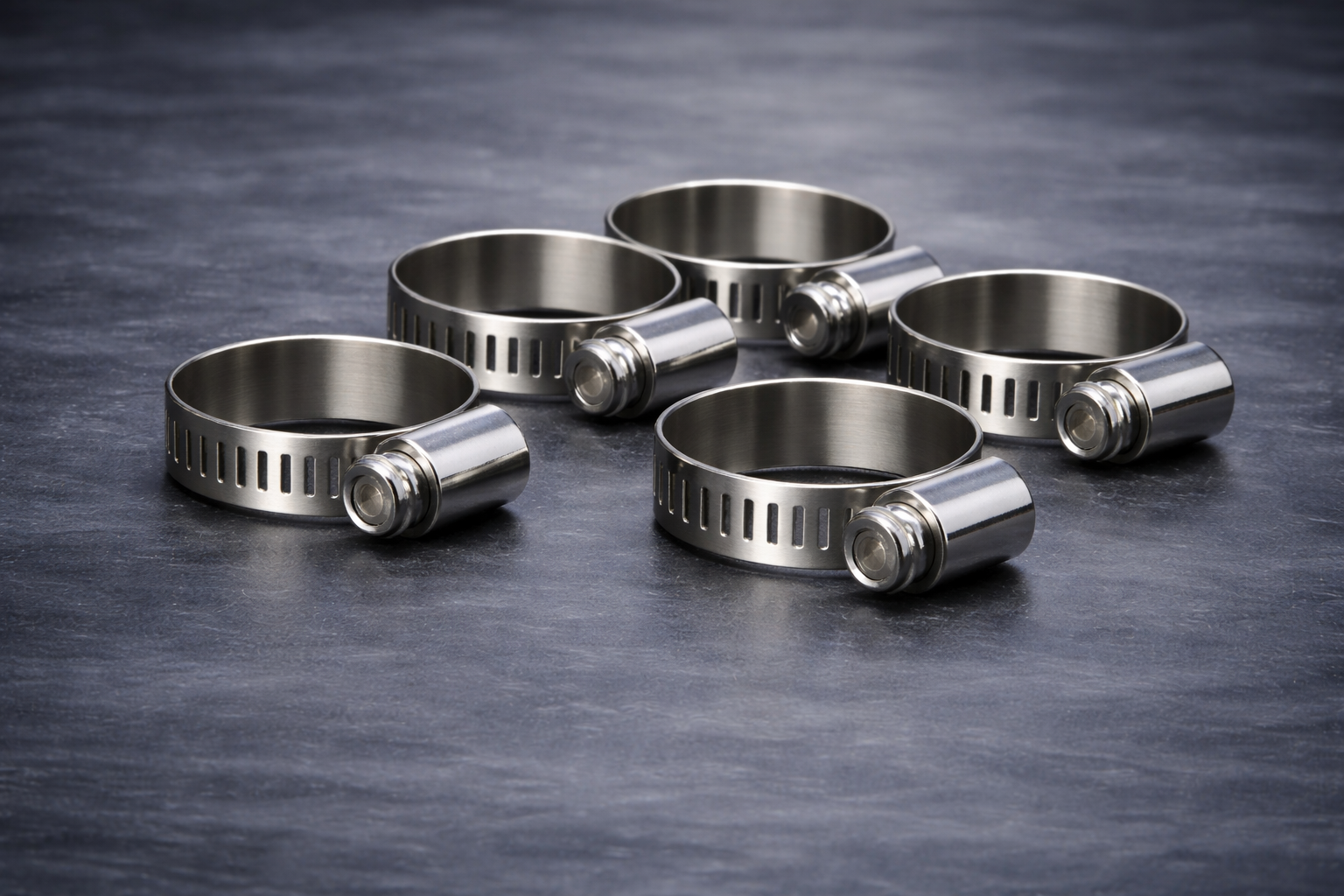 Five metal hose clamps with screw bands lying on a dark textured surface.
