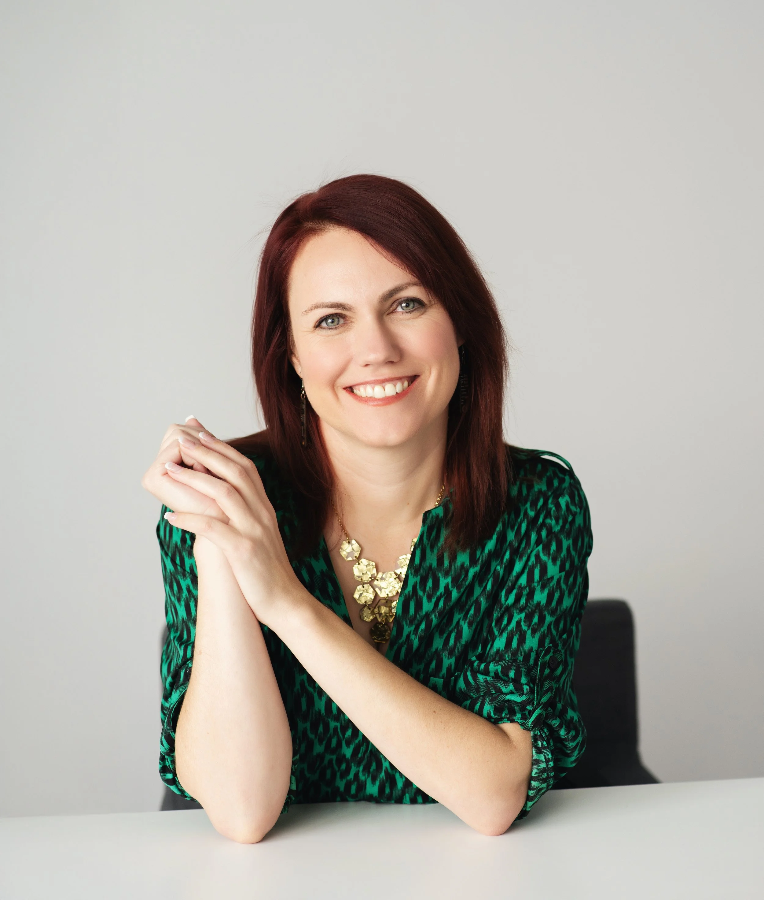 Katrina German with red hair smiling, wearing a green patterned blouse and a gold necklace, sitting at a table against a plain light gray background.